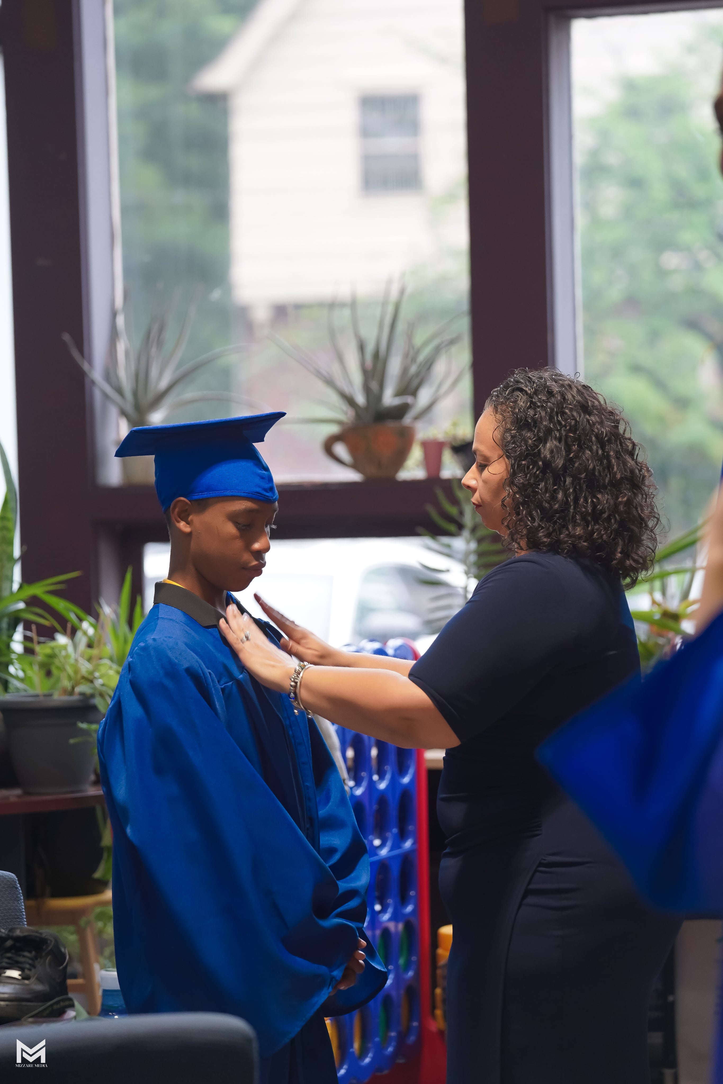 A woman adjusting the cap and gown of a boy in a blue graduation outfit, inside a bright room with plants visible in the background.