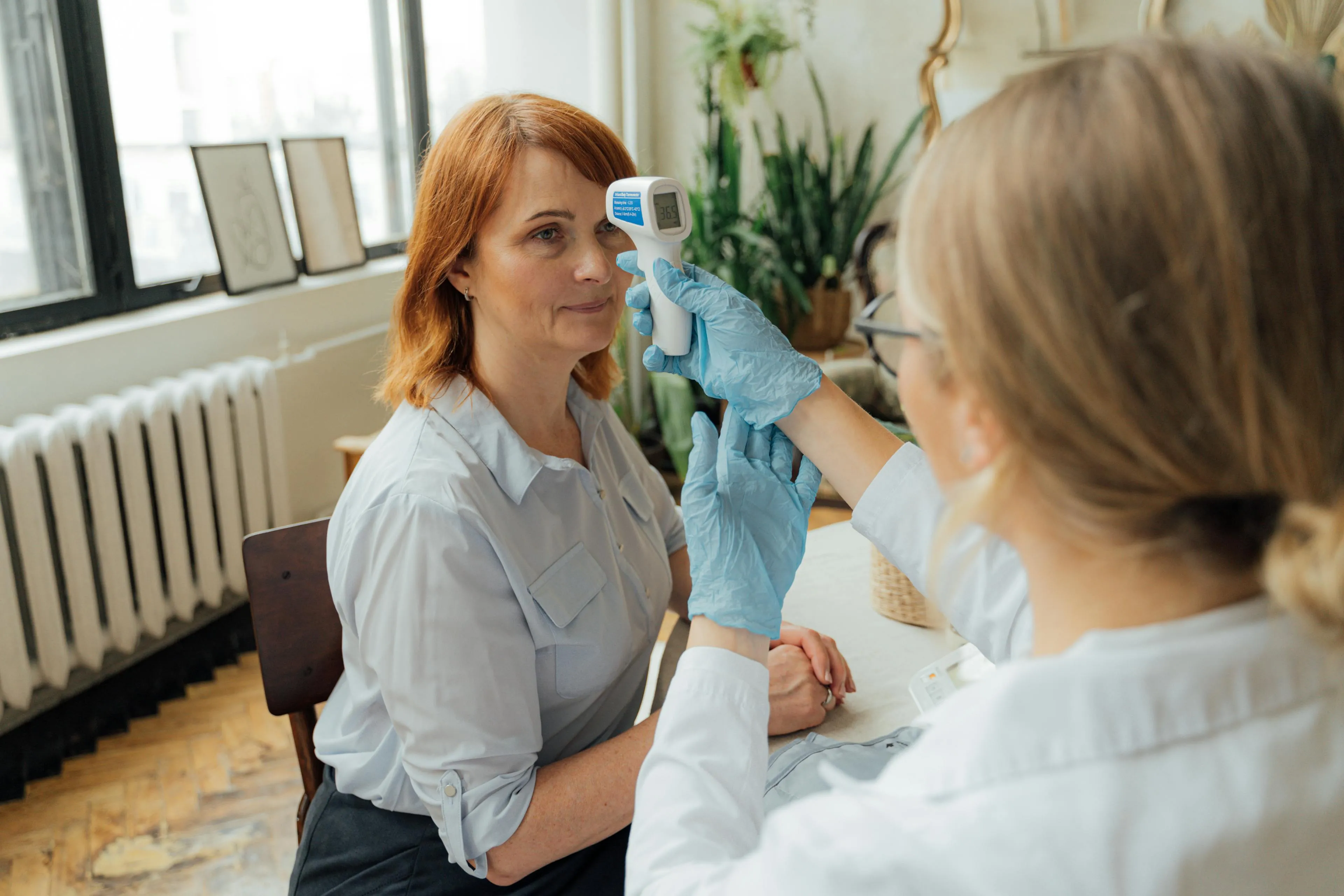 Healthcare professional wearing blue gloves checks a woman's forehead temperature with a digital infrared thermometer indoors.