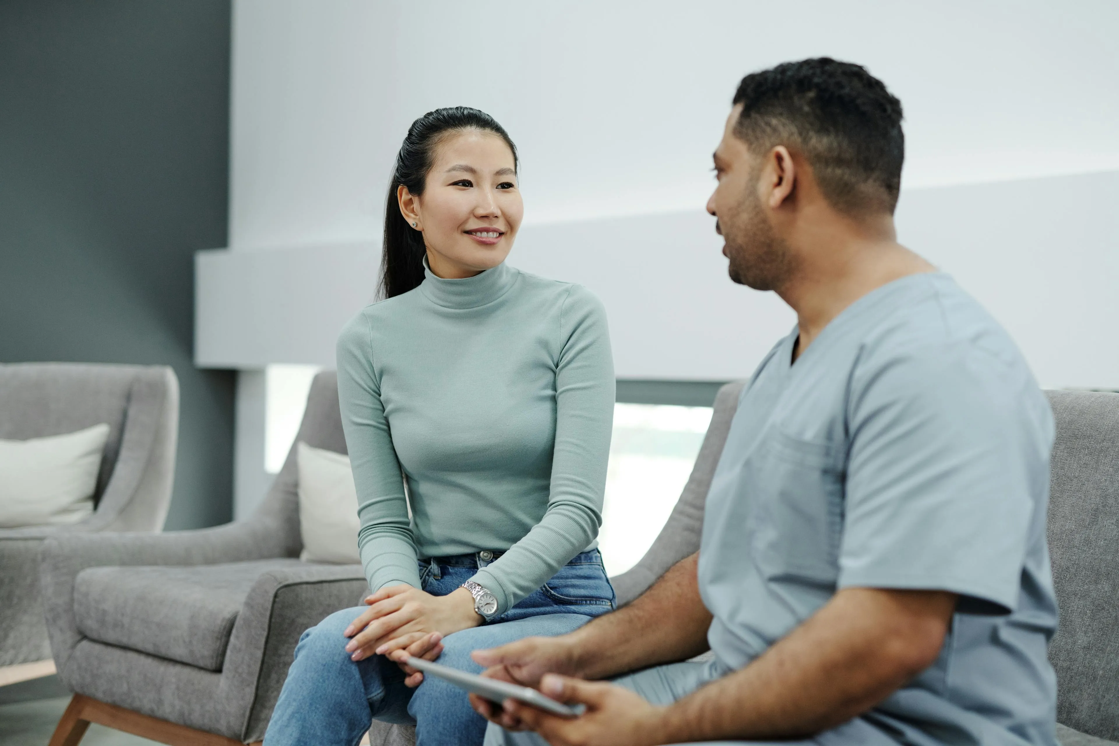 A woman in a green turtleneck and jeans talking to a man in blue medical scrubs sitting on gray chairs in a modern waiting area.
