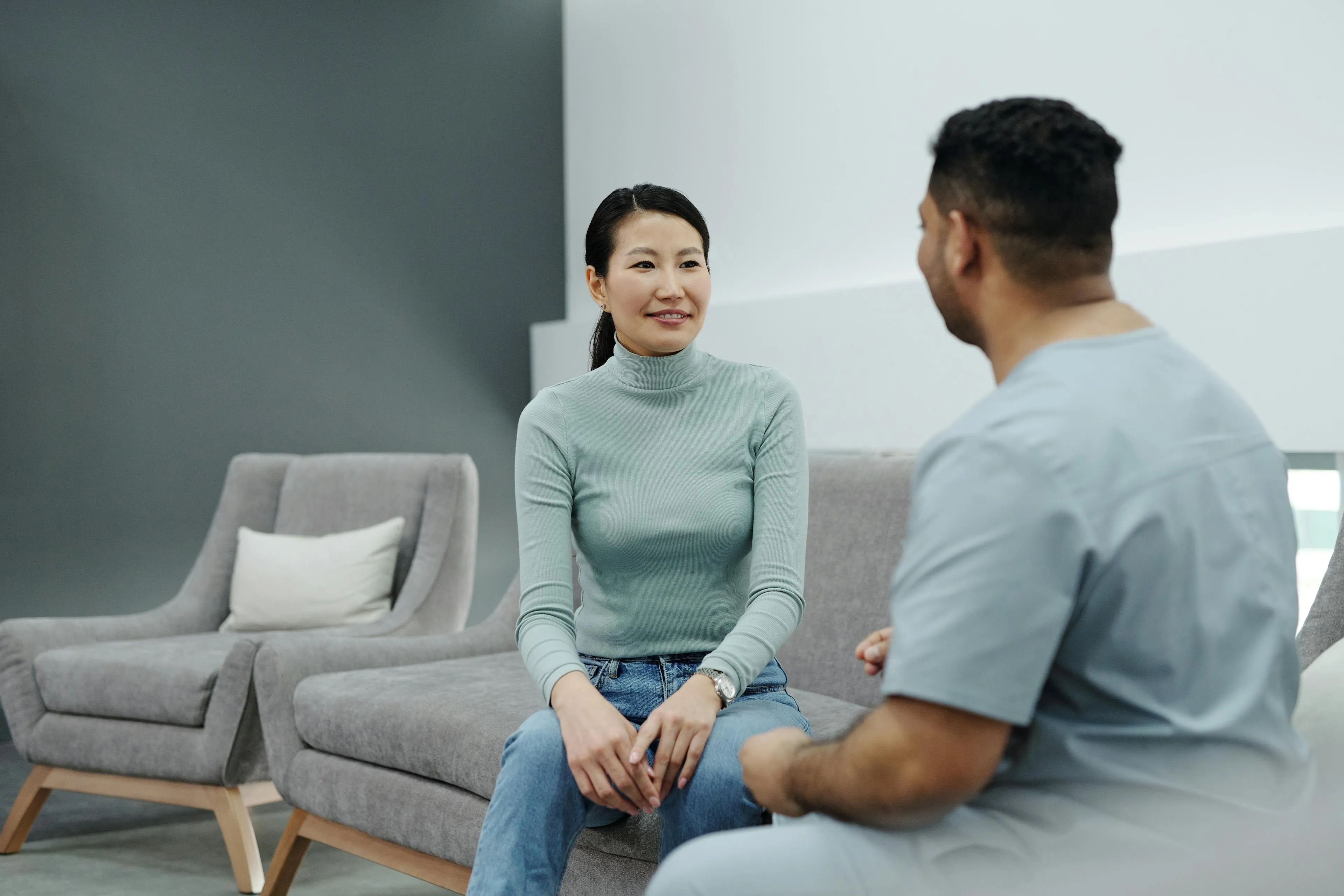 Woman in a green turtleneck sitting on a gray sofa facing a man in light blue scrubs in a modern room.