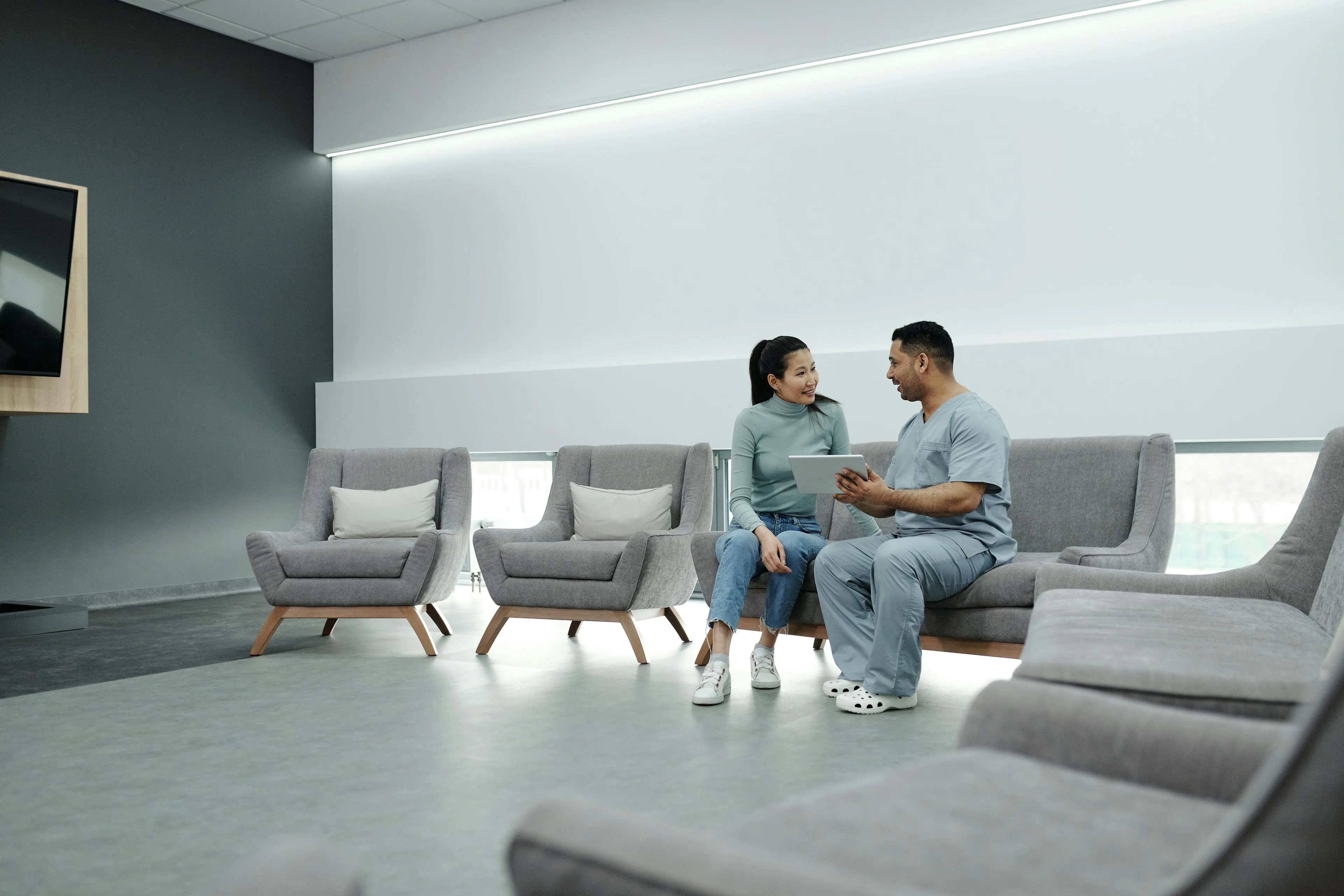 Healthcare professional in scrubs showing a tablet to a woman sitting together in a modern waiting area with gray armchairs.