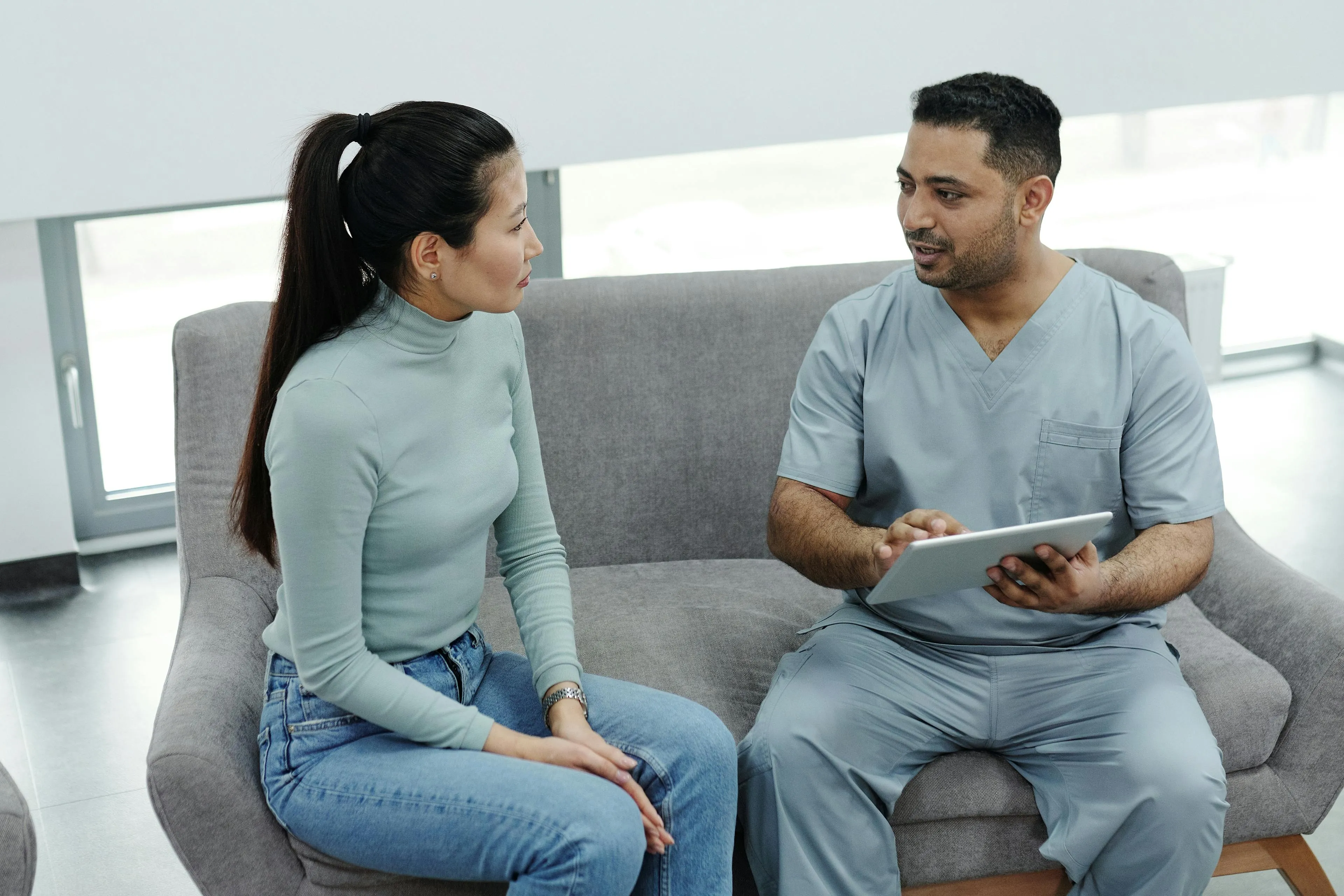 A healthcare professional in scrubs talks with a seated woman wearing a light green turtleneck and jeans in a bright room.