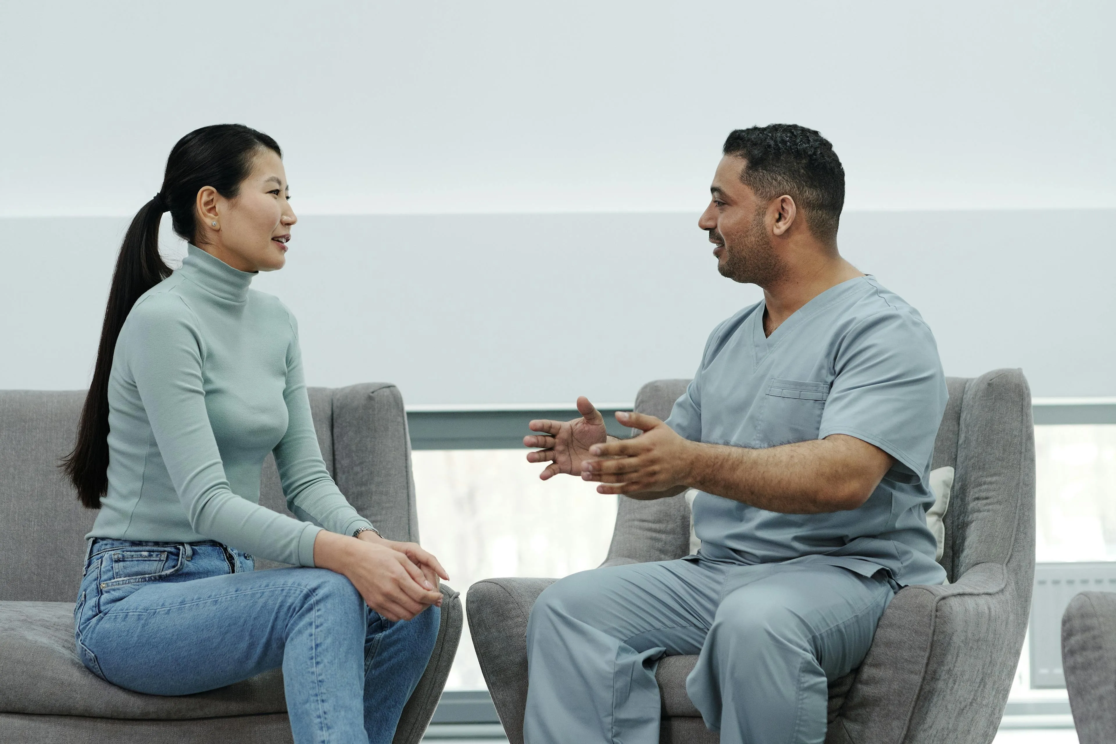 A male healthcare professional in scrubs talking with a seated woman wearing a light blue turtleneck and jeans in a bright room.