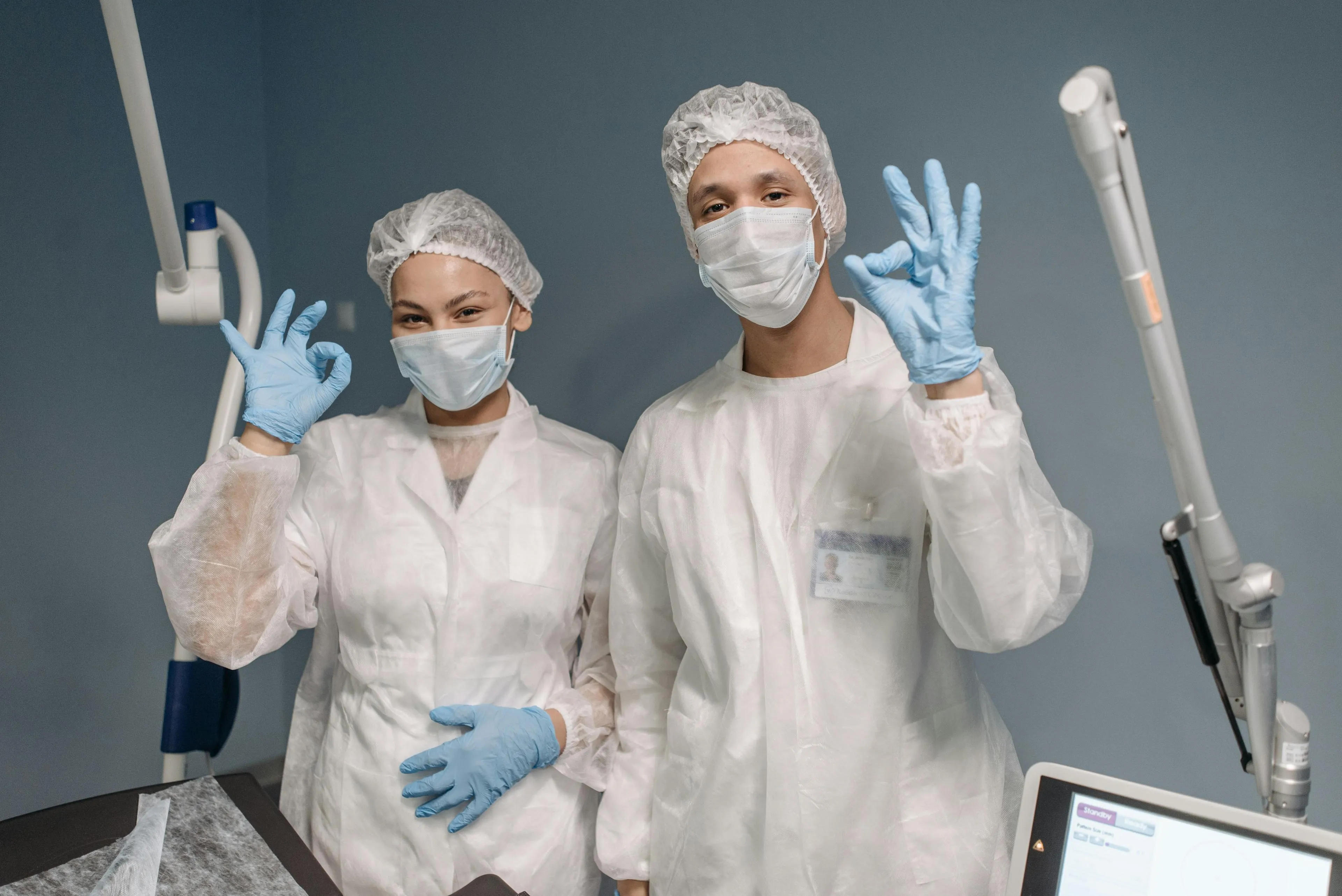 Two healthcare workers in white protective gowns, masks, hairnets, and blue gloves showing OK hand signs in a medical setting.