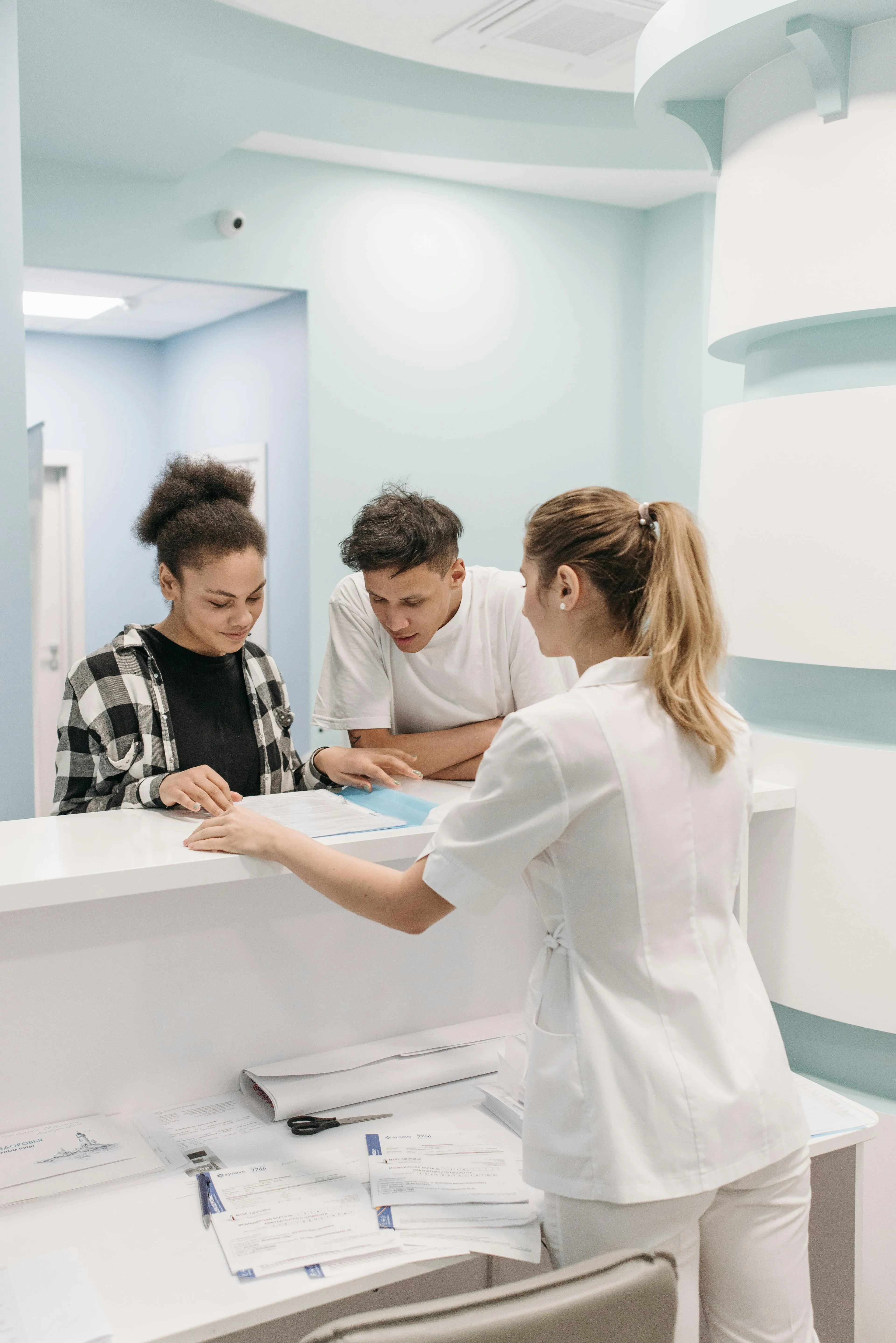 Healthcare professional in white uniform assisting two patients at a reception desk with documents.