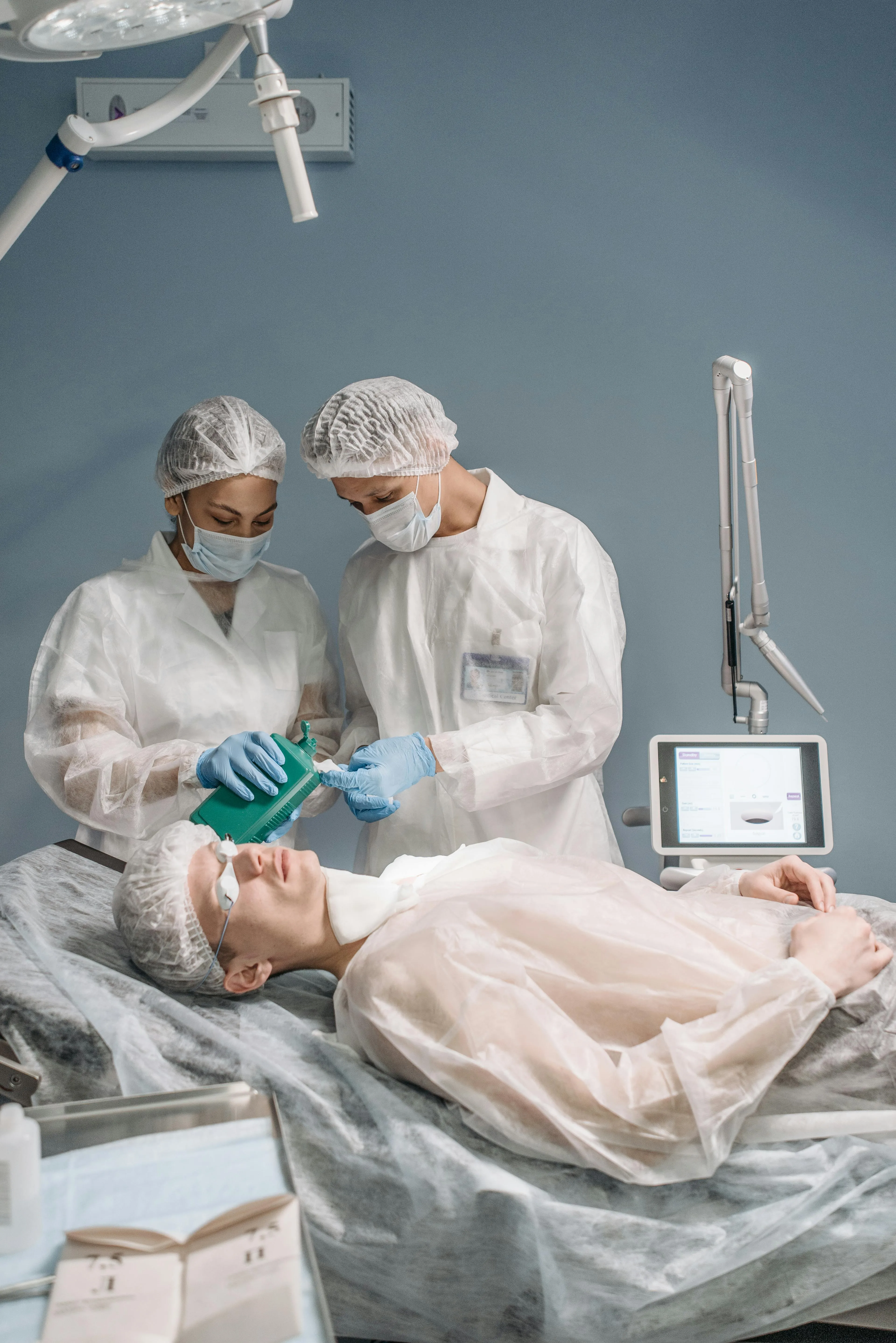 Two medical professionals in protective gear preparing a patient lying on a treatment table for a procedure, with medical equipment nearby.