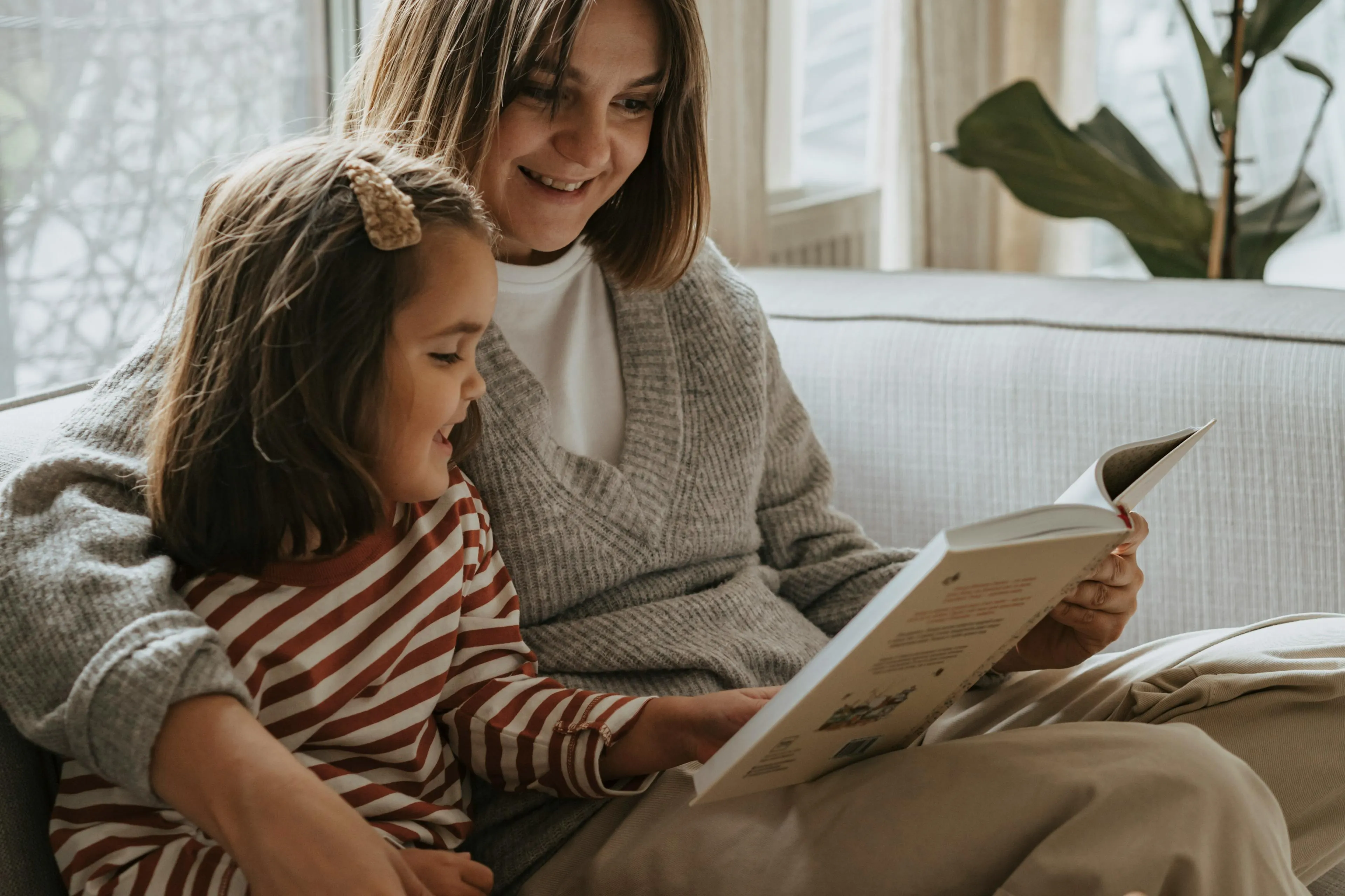 Smiling woman in gray sweater reading a book with a young girl on a couch.