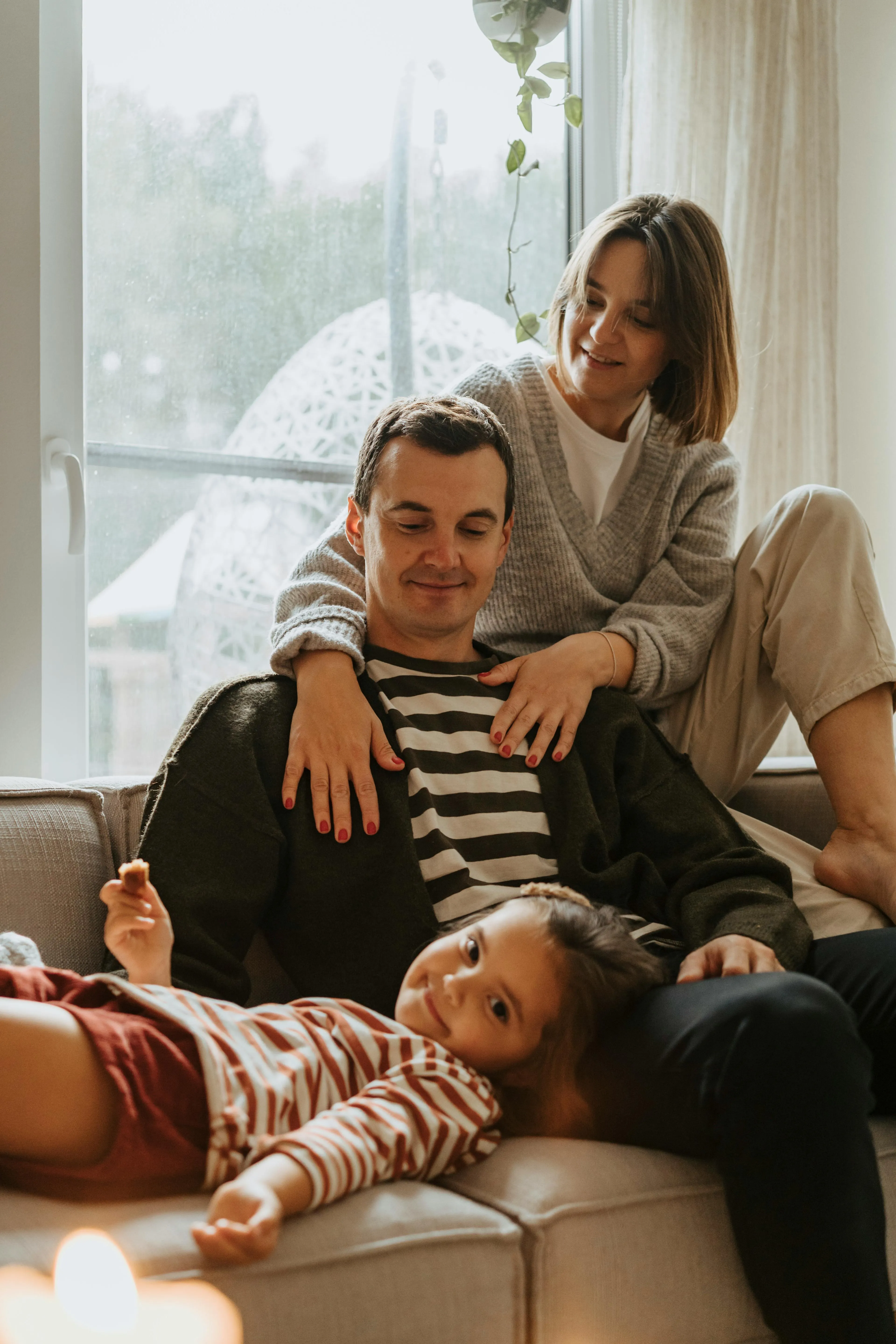 Smiling family of three relaxing on a couch near a window, with the child lying across the father's lap.