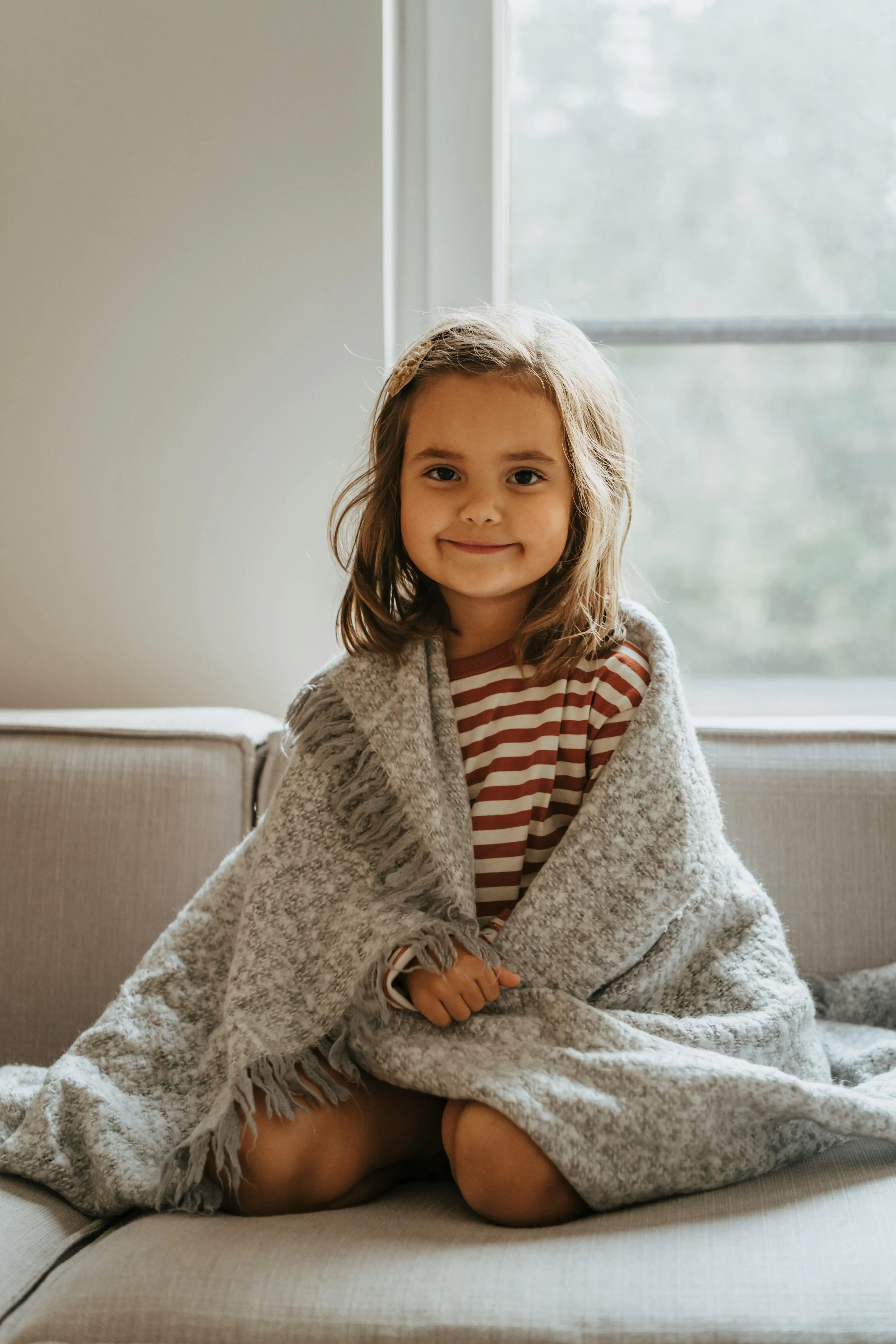 Smiling young girl sitting on a couch wrapped in a gray fringed blanket with a window in the background.