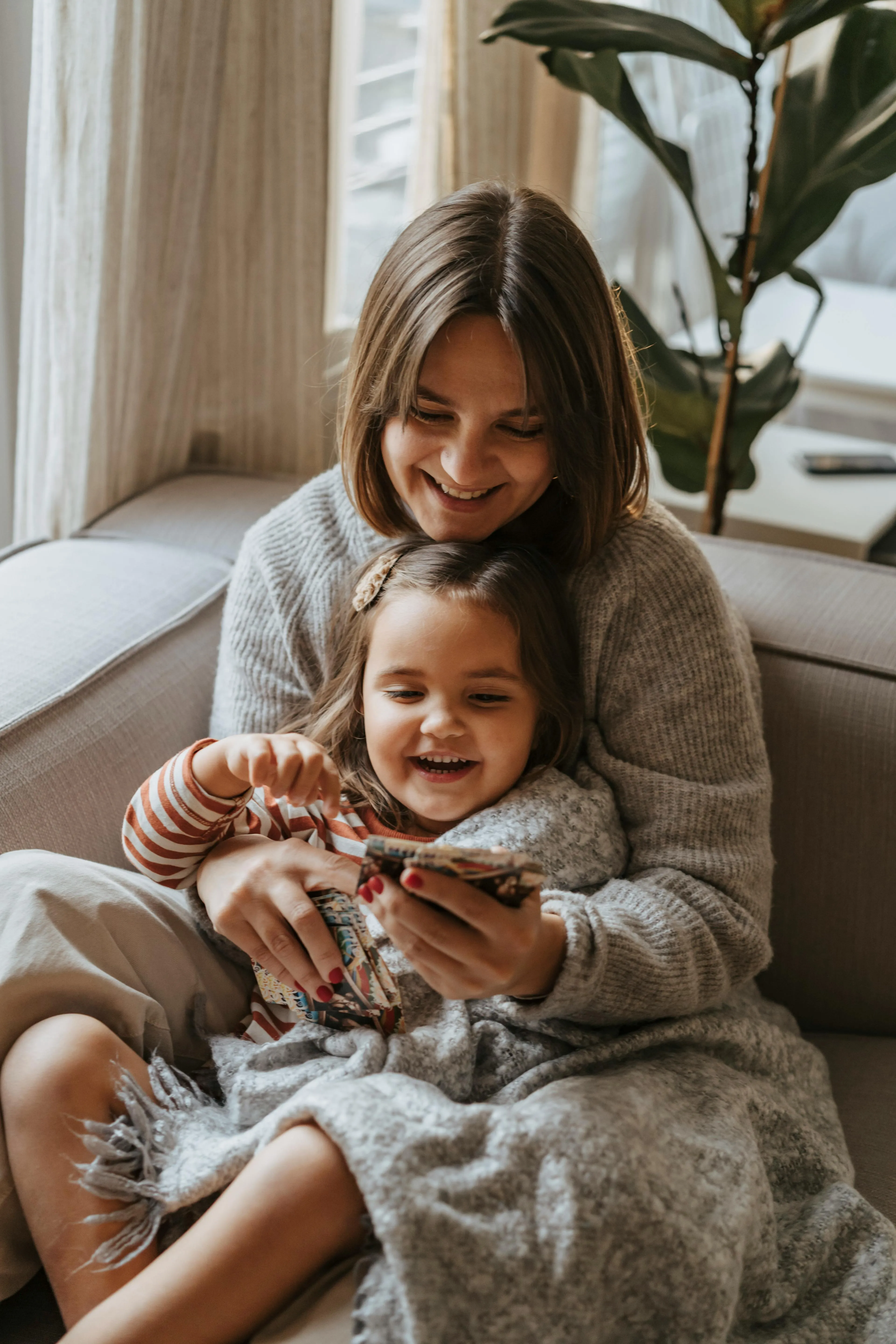 Mother and daughter smiling and looking at a smartphone while sitting on a couch wrapped in a gray blanket.