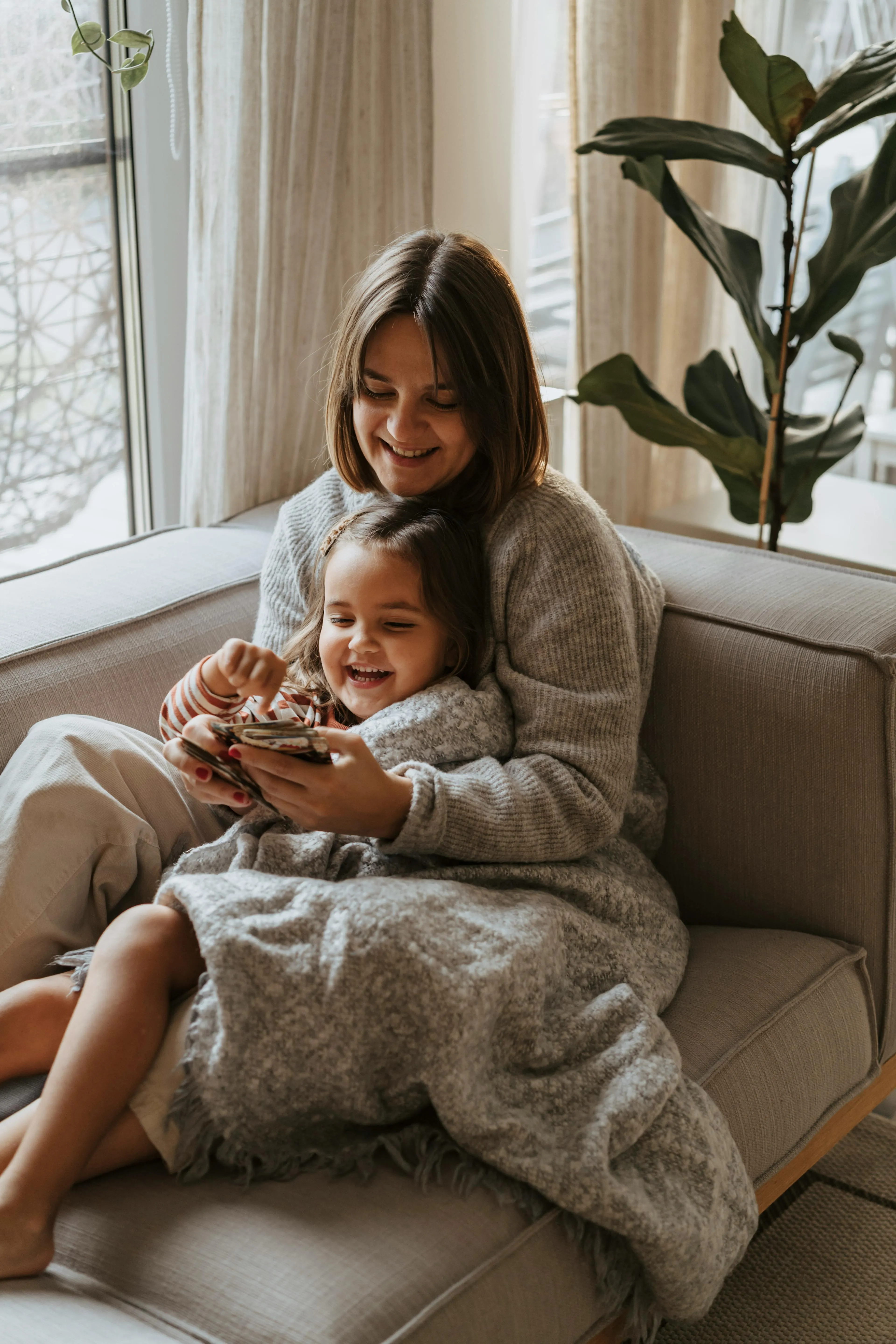 Mother and daughter sitting on a couch wrapped in a gray blanket, smiling and looking at a smartphone together.