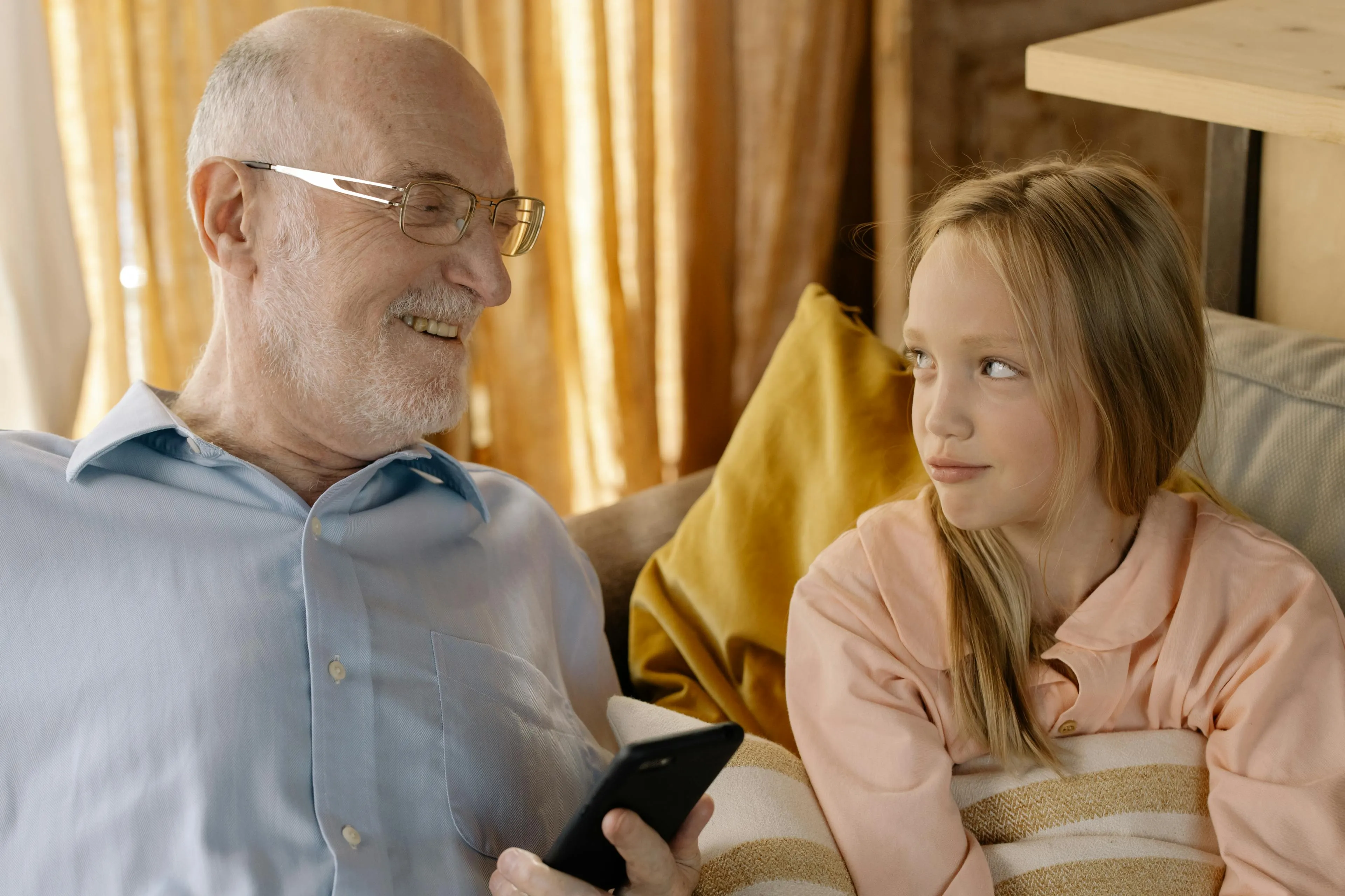 Smiling elderly man holding a smartphone looks at a girl sitting beside him on a couch.