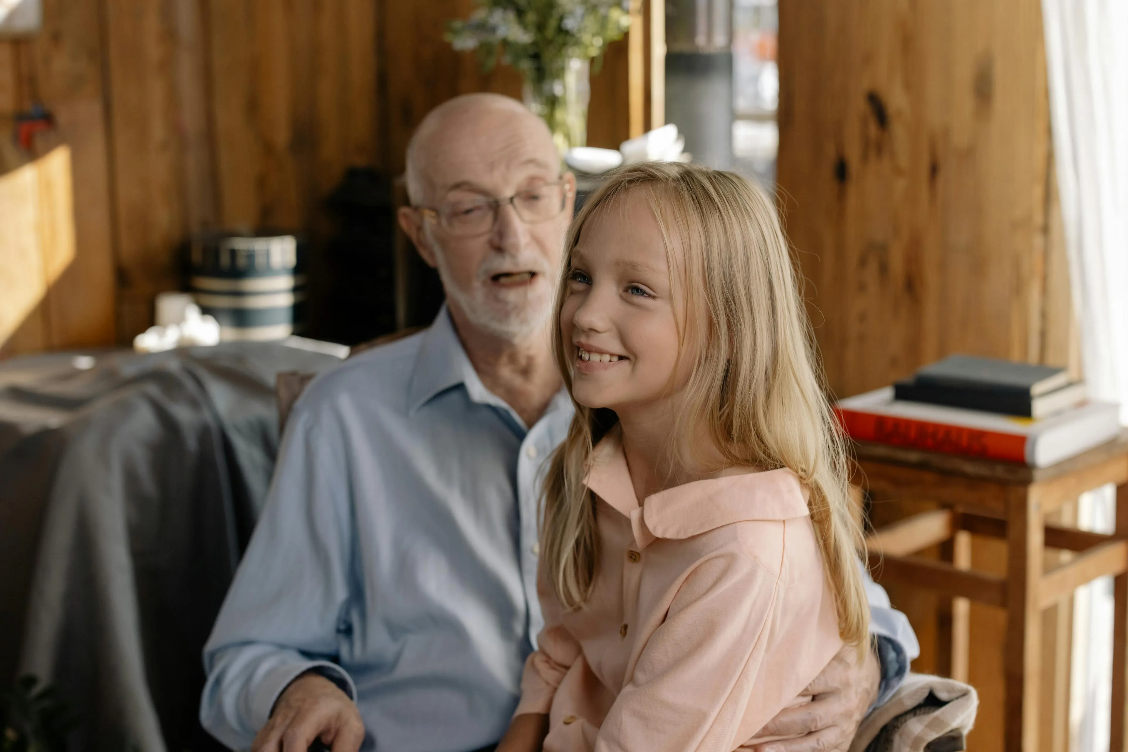 Smiling elderly man in glasses holding a cheerful young girl with long blonde hair indoors.