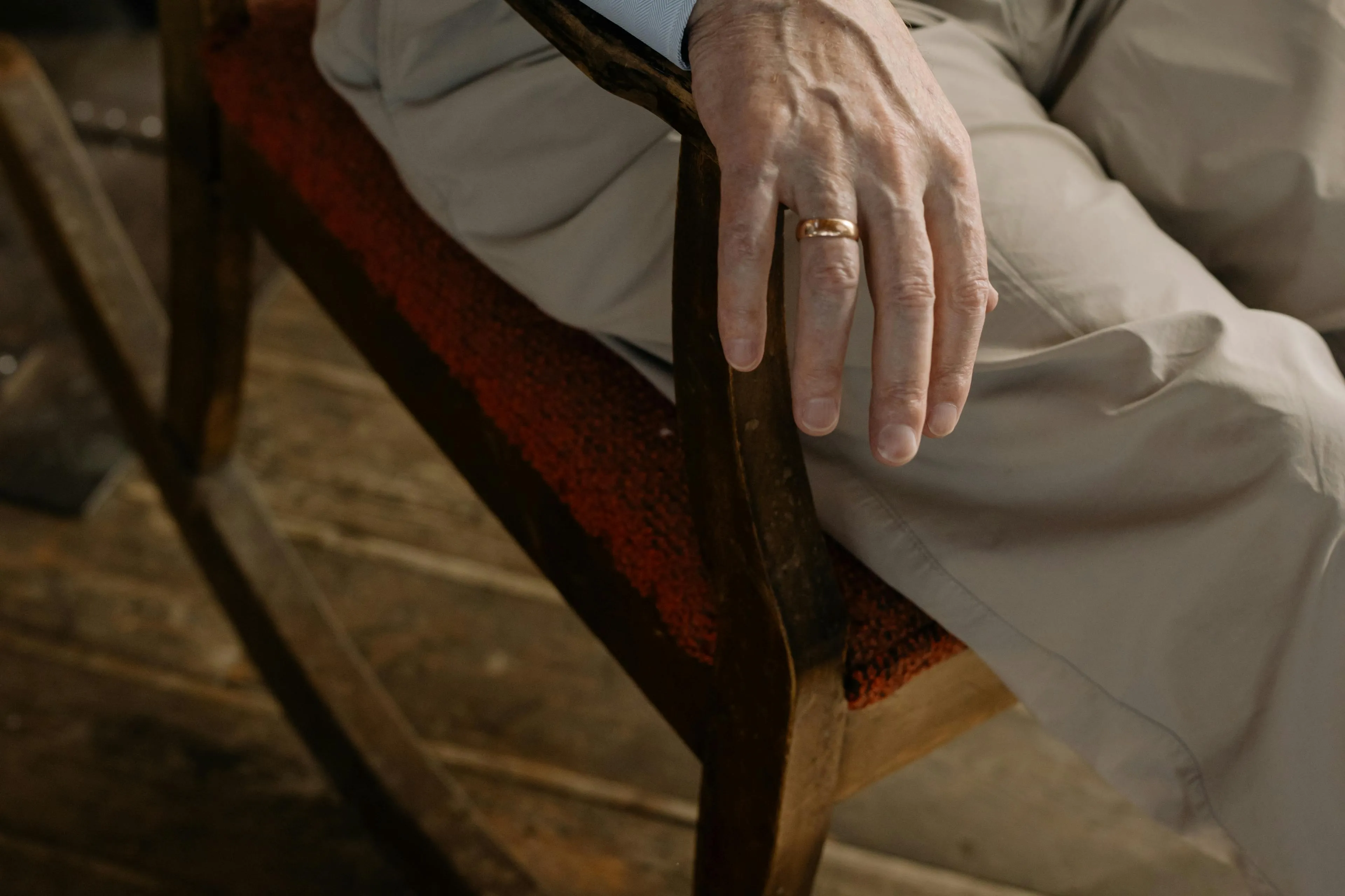 Close-up of an elderly person's hand with a wedding ring resting on the arm of a wooden rocking chair with a red cushion.