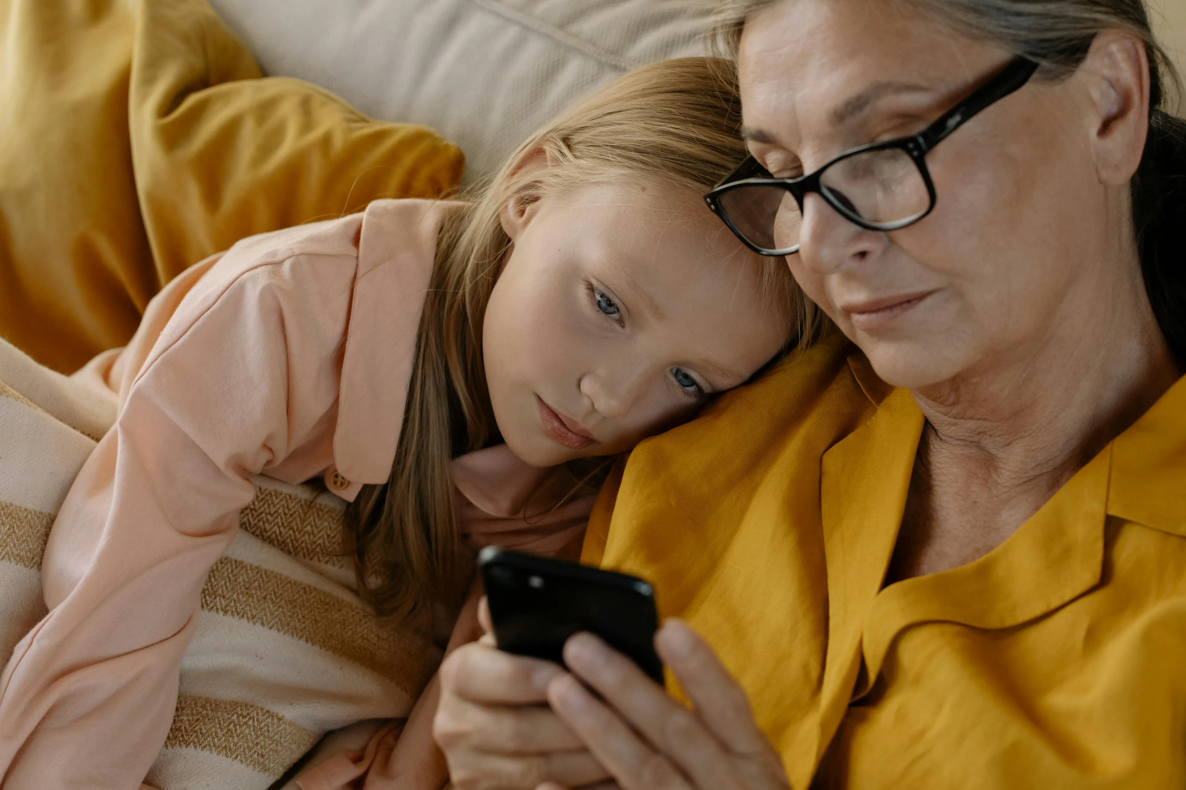 Elder woman wearing glasses and a mustard yellow shirt looking at a smartphone while a young girl rests her head on the woman's shoulder.