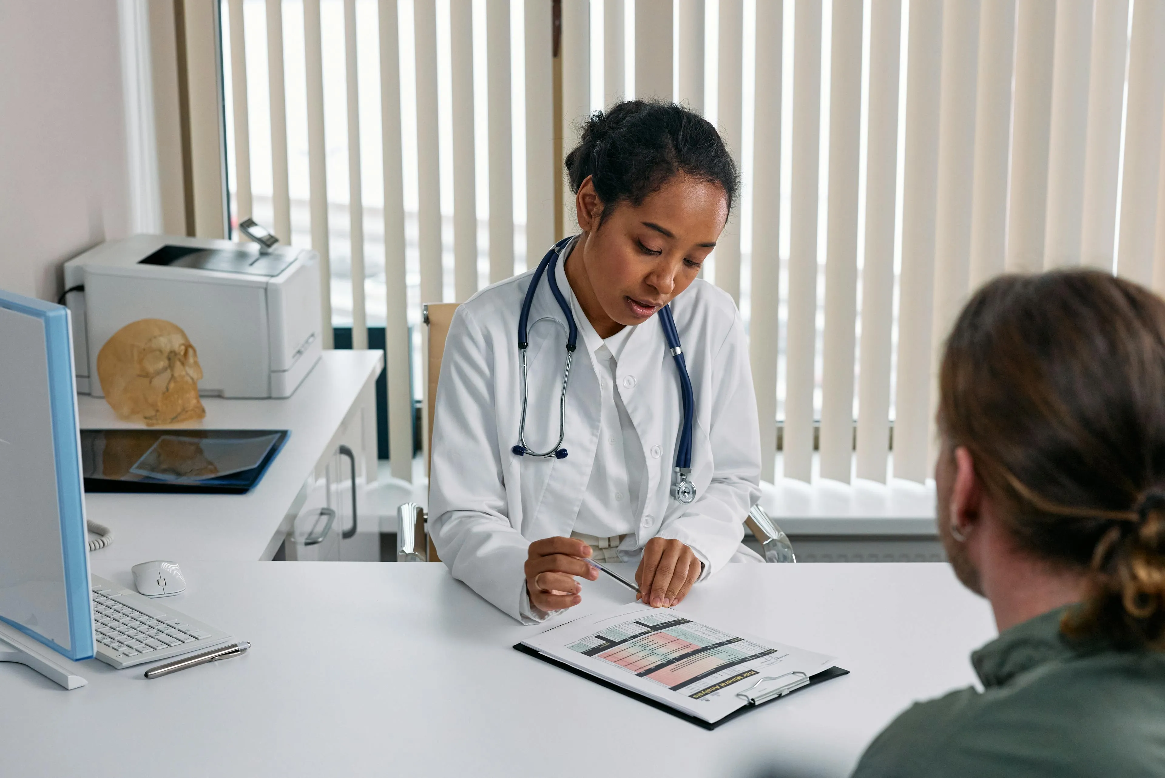 Female doctor in white coat with stethoscope explaining a medical chart to a seated patient in a consultation room.