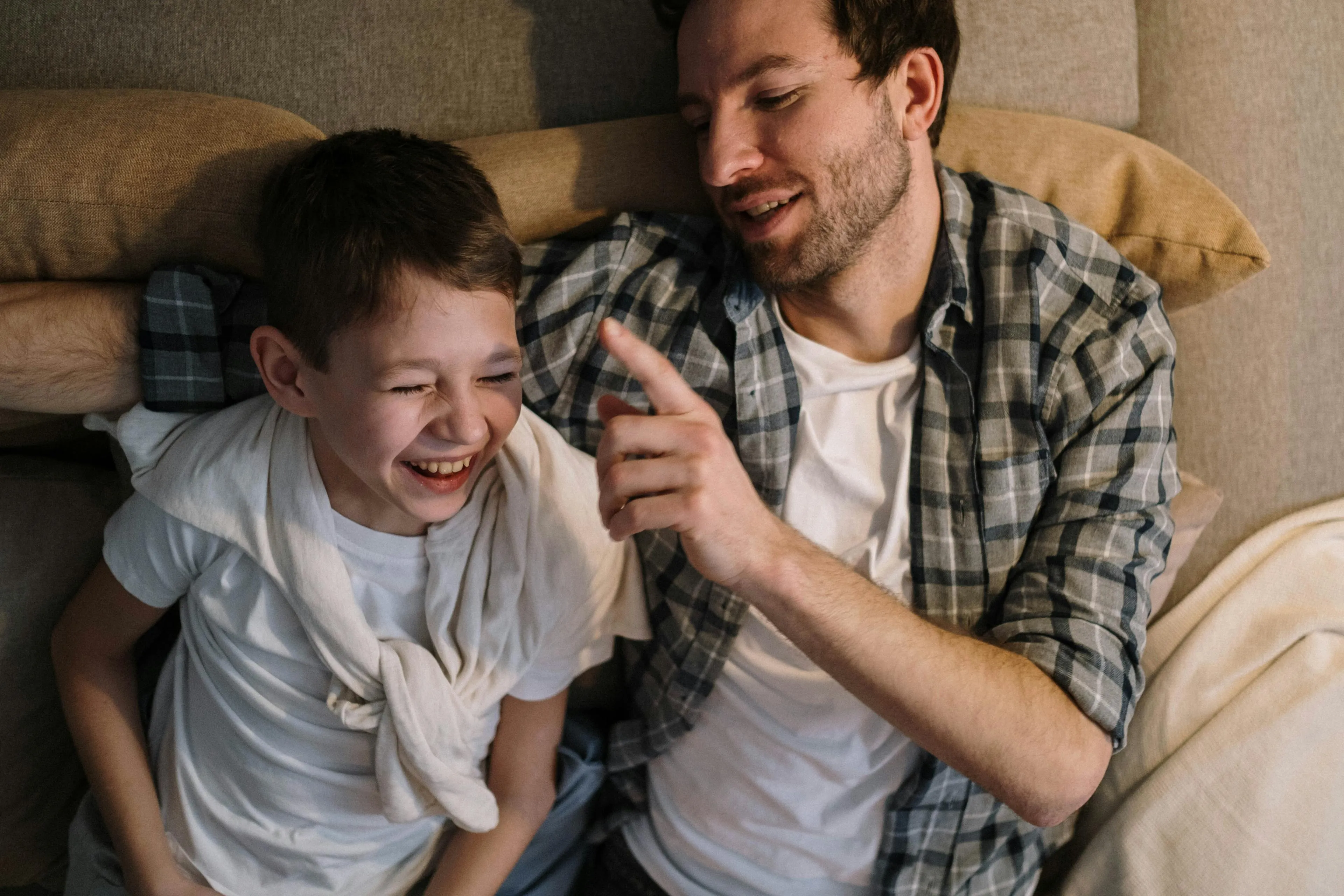 Man and boy lying on a couch, the man playfully pointing finger while the boy laughs with eyes closed.