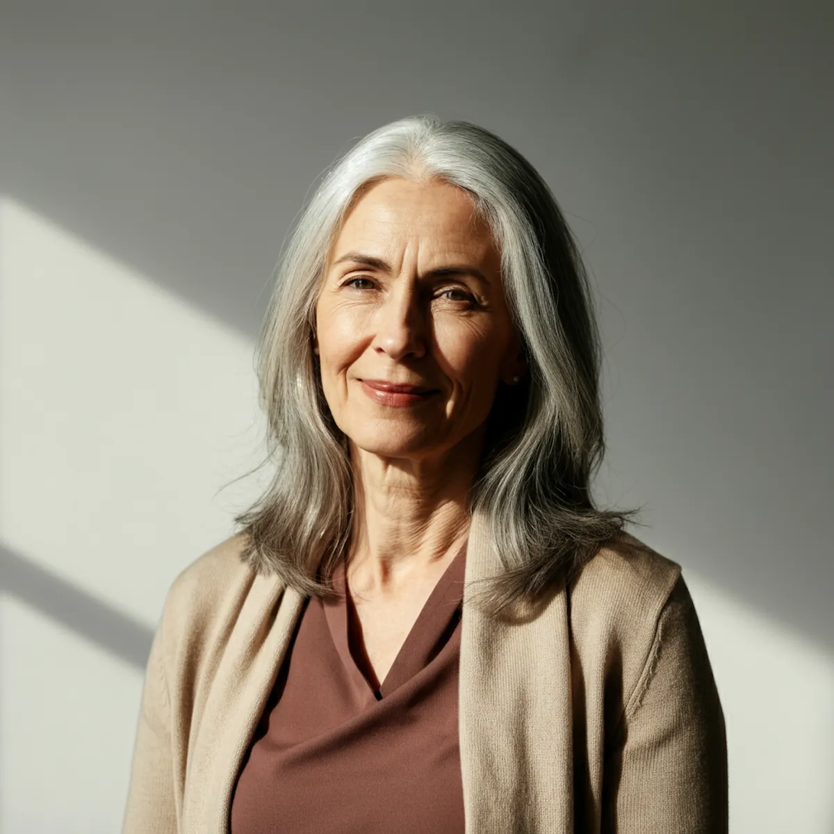 Portrait of a serene older woman with silver hair wearing a beige cardigan and brown top against a light gray background.