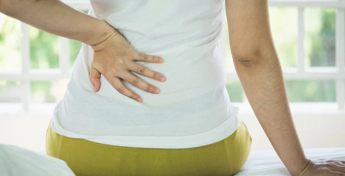 Person in a white shirt holding their lower back while seated on a bed