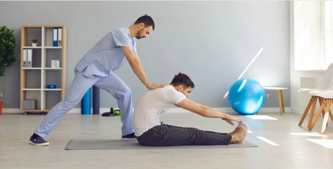 Trainer assisting a seated man with a forward stretch on a mat.