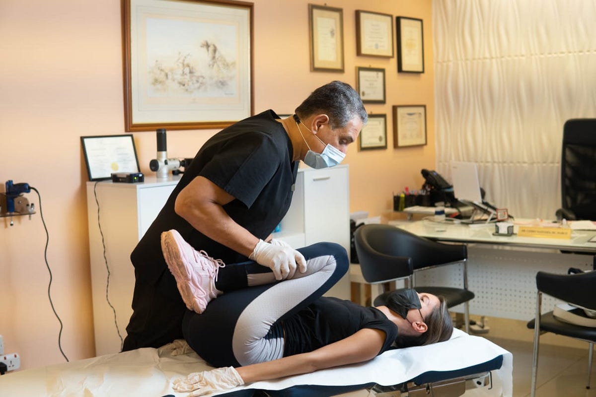 Masked practitioner guides a woman through a leg stretch on a treatment table.