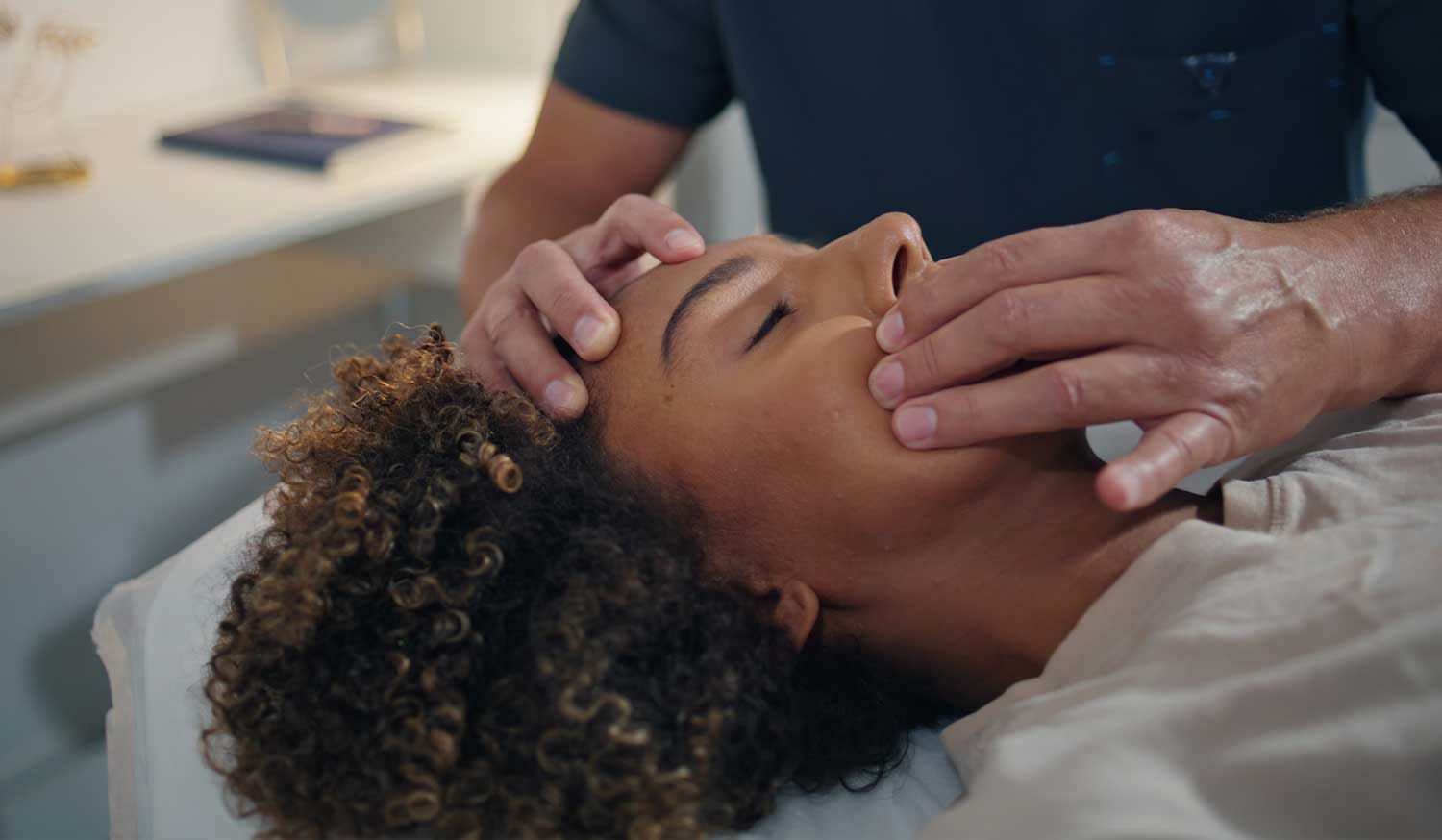 Practitioner gently supports a person’s jaw and head while they lie on a treatment table.