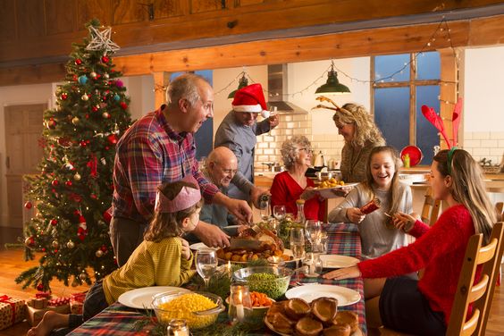 Family enjoying a Christmas dinner beside a decorated tree, with a child in a Santa hat and festive table setting