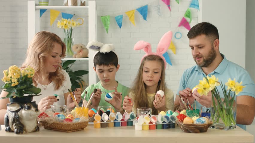 Family decorating Easter eggs at a table with paint, baskets, flowers, and bunny decorations.