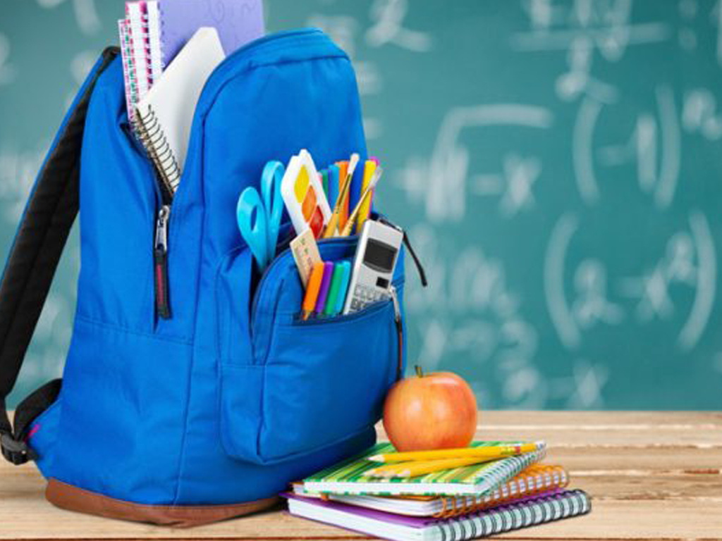 Blue backpack filled with school supplies beside an apple and stacked notebooks, in front of a chalkboard.