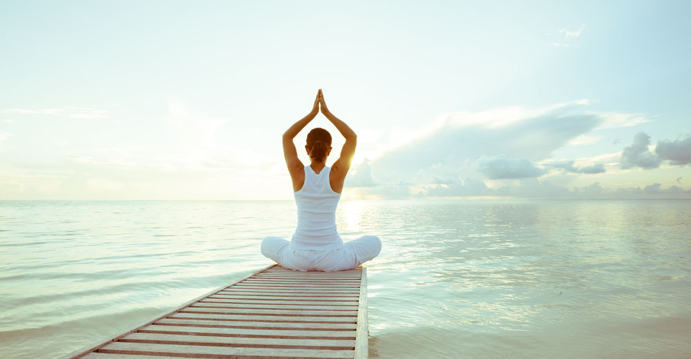 Person sitting cross-legged in a yoga pose on a dock over calm water at sunrise
