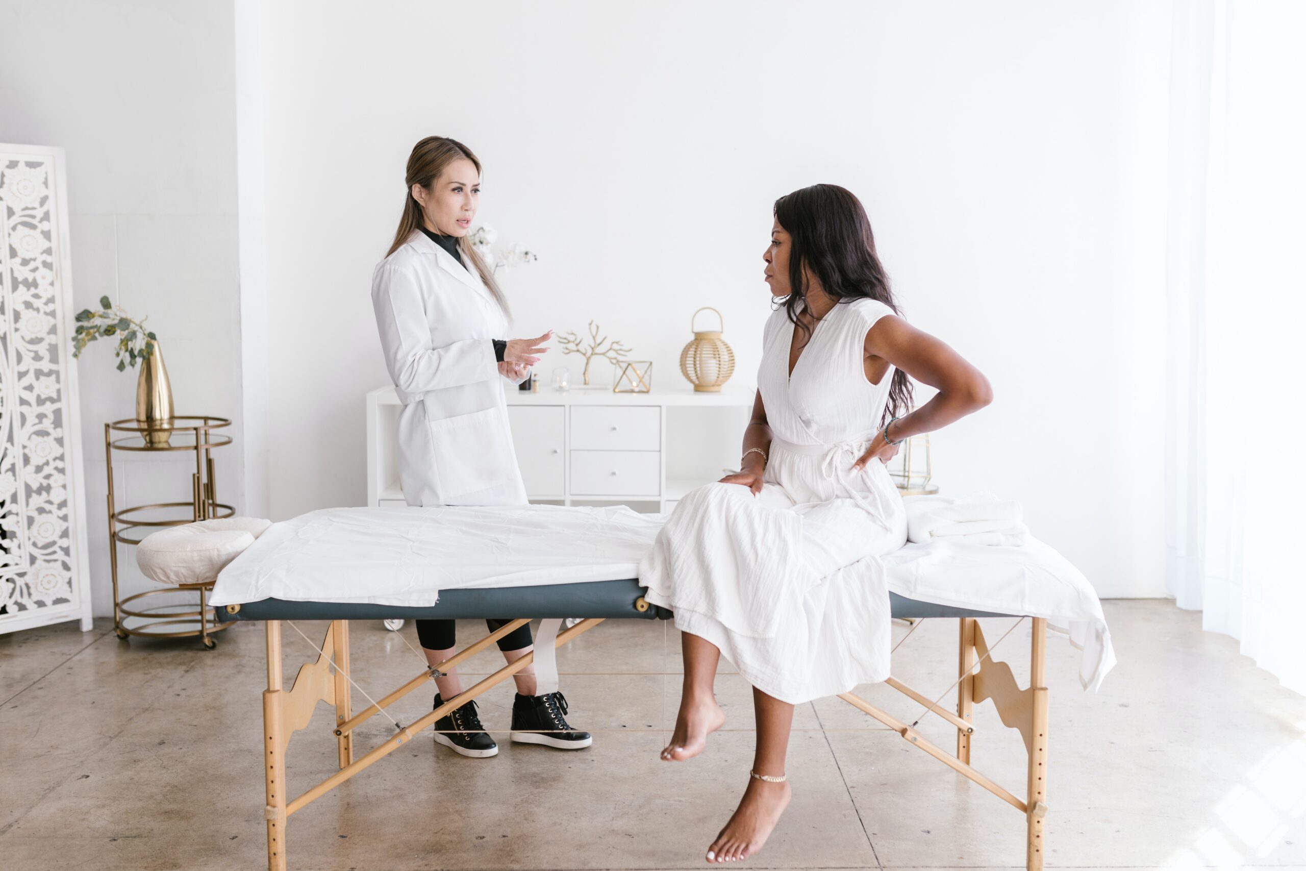Healthcare professional speaks with a seated woman holding her lower back in a treatment room.