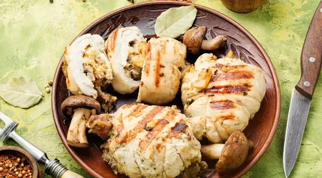Dans un plat creux se trouve les morceaux de poulet grillés ainsi que des champignons entiers. Une ballotine coupée permet de voir la farce.