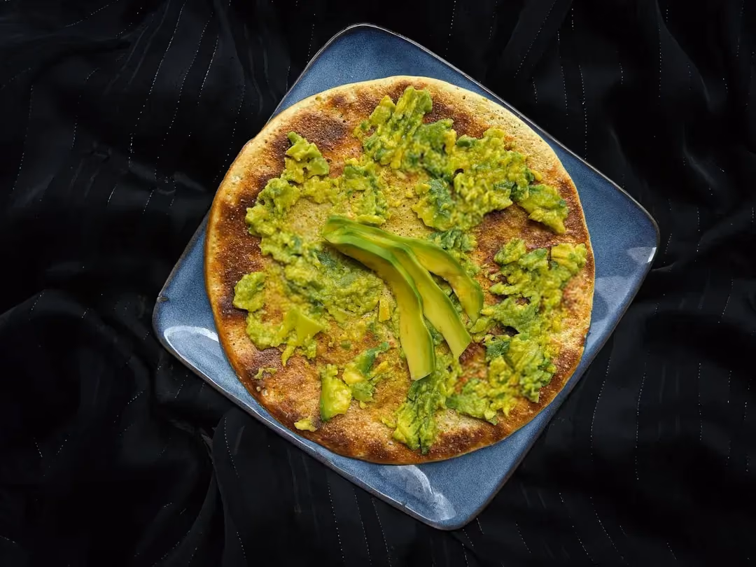 A shallow square dish with rounded edges is used to present a big pancake topped with avocado.