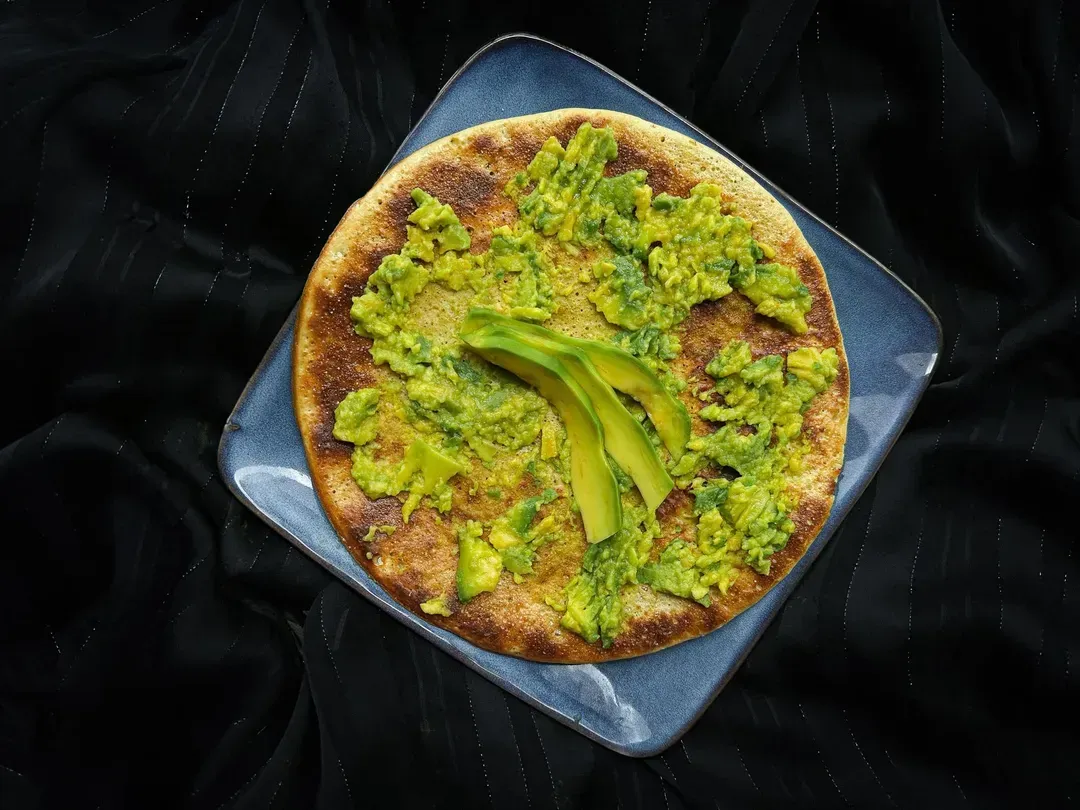 A shallow square dish with rounded edges is used to present a big pancake topped with avocado.