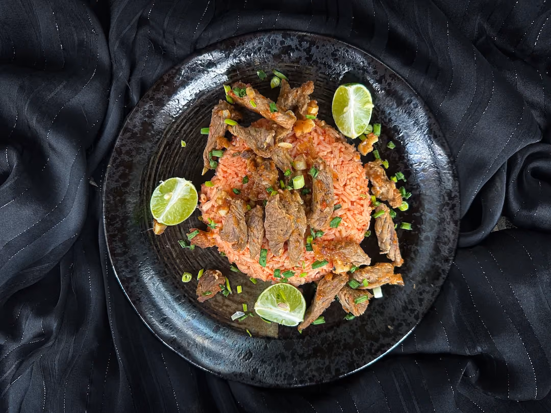 Le riz coloré par la sauce tomate est recouvert par le faux-filet de boeuf. Un citron vert coupé en quatre est disposé autour du plat.