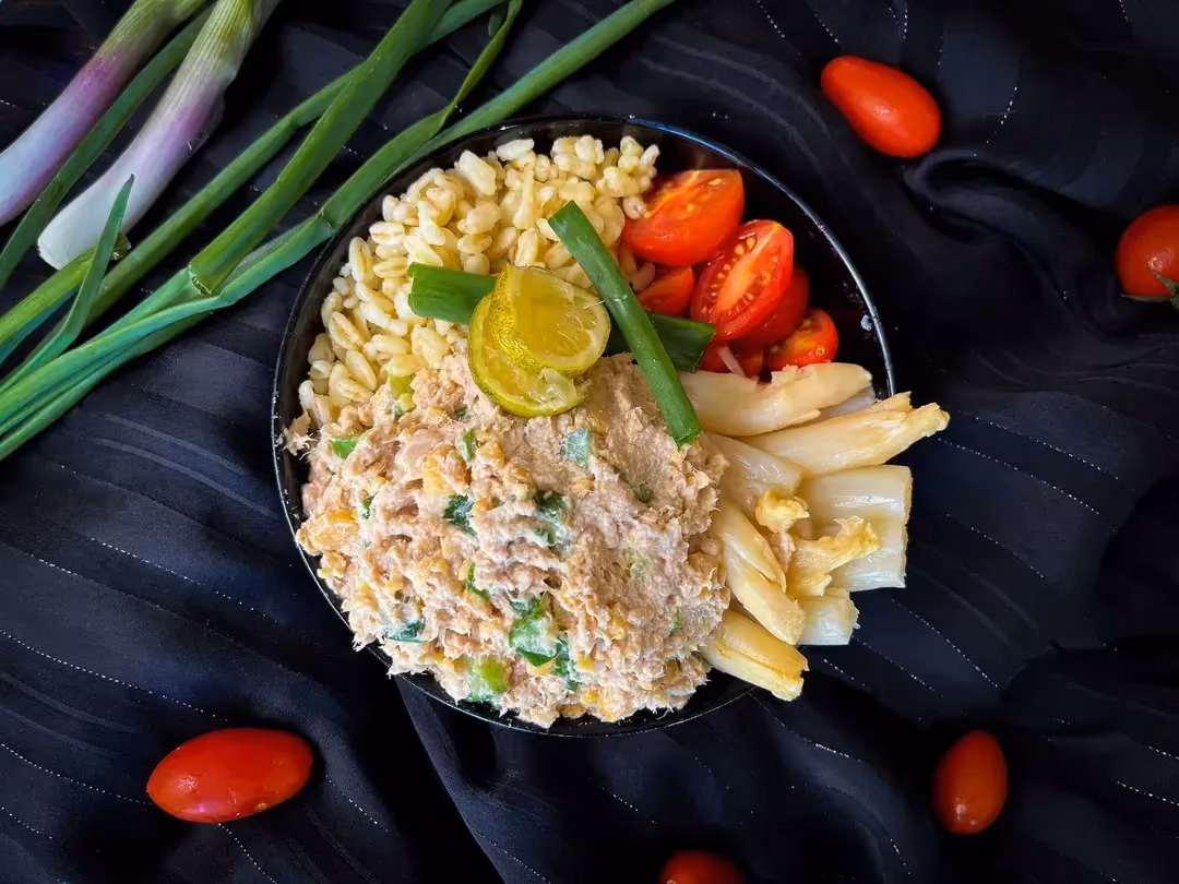 In a small bowl, there is wheat with asparagus tips, cherry tomatoes cut in half, and tuna mixed with skyr.