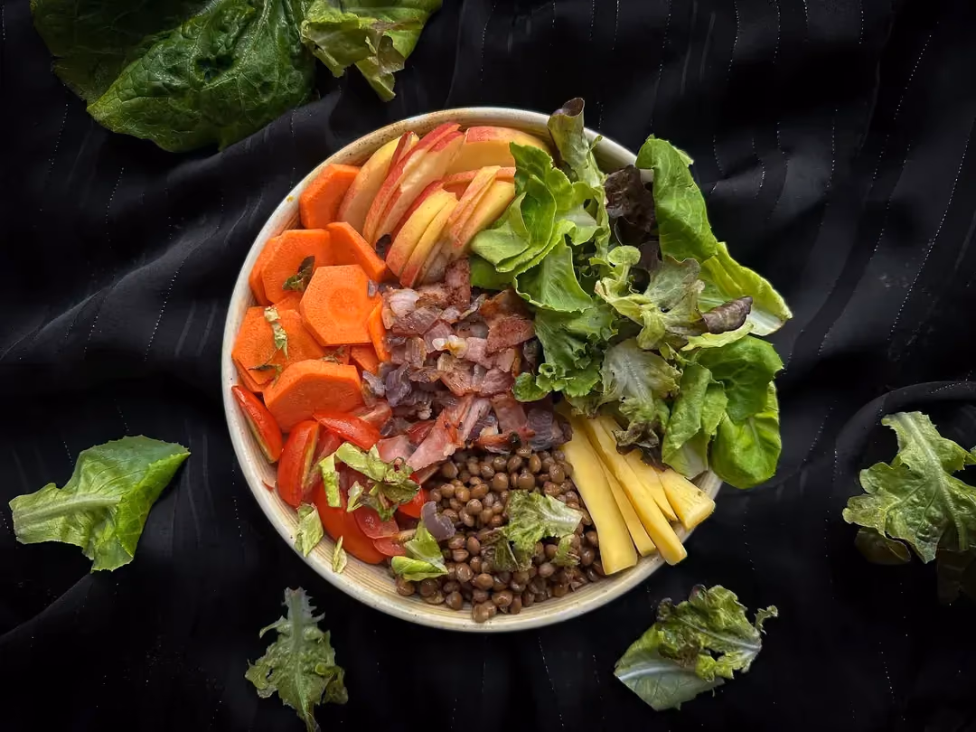 In a stoneware bowl are all the ingredients for the recipe: lentils, bacon lardons, Comté cheese, and raw vegetables.