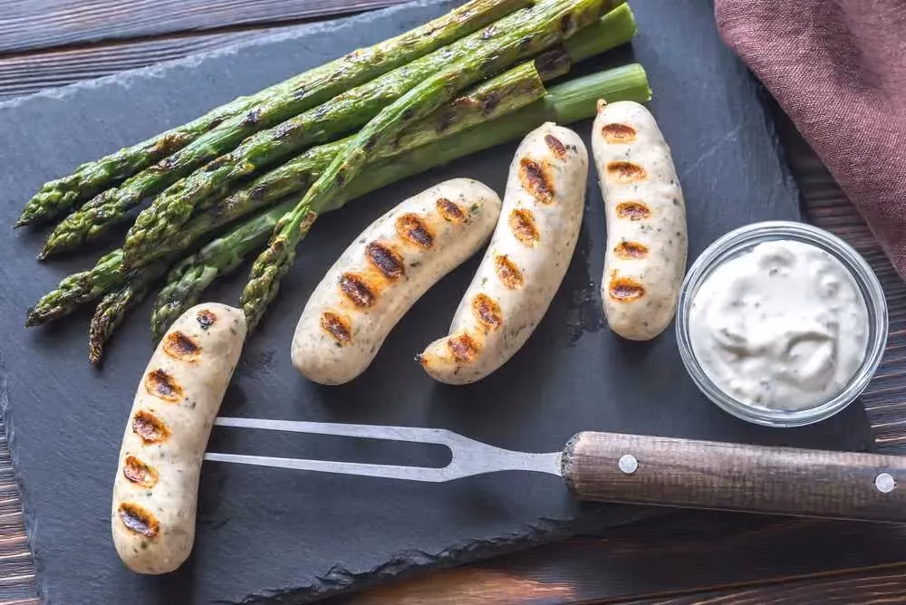 Quatre boudins blancs grillés dont l'un est embroché repose sur une plaque de cuisson. Quelques asperges sont déposées à côté.