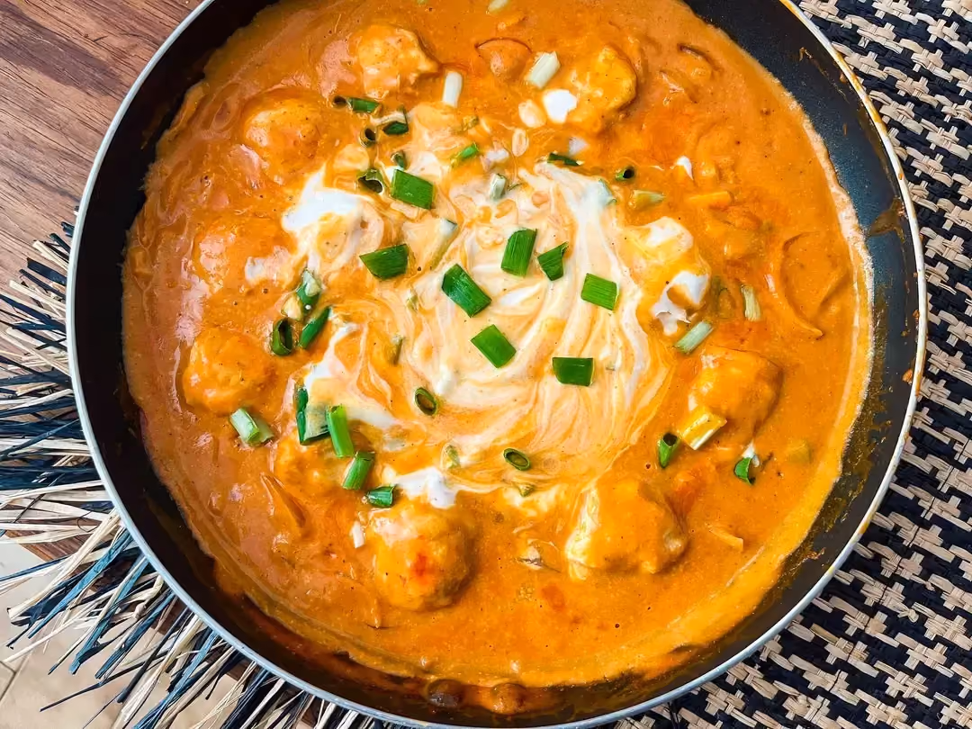 Posée sur une table en bois, se trouve une poêle contenant les boulettes de poulet, le coulis de tomates et le lait de coco.