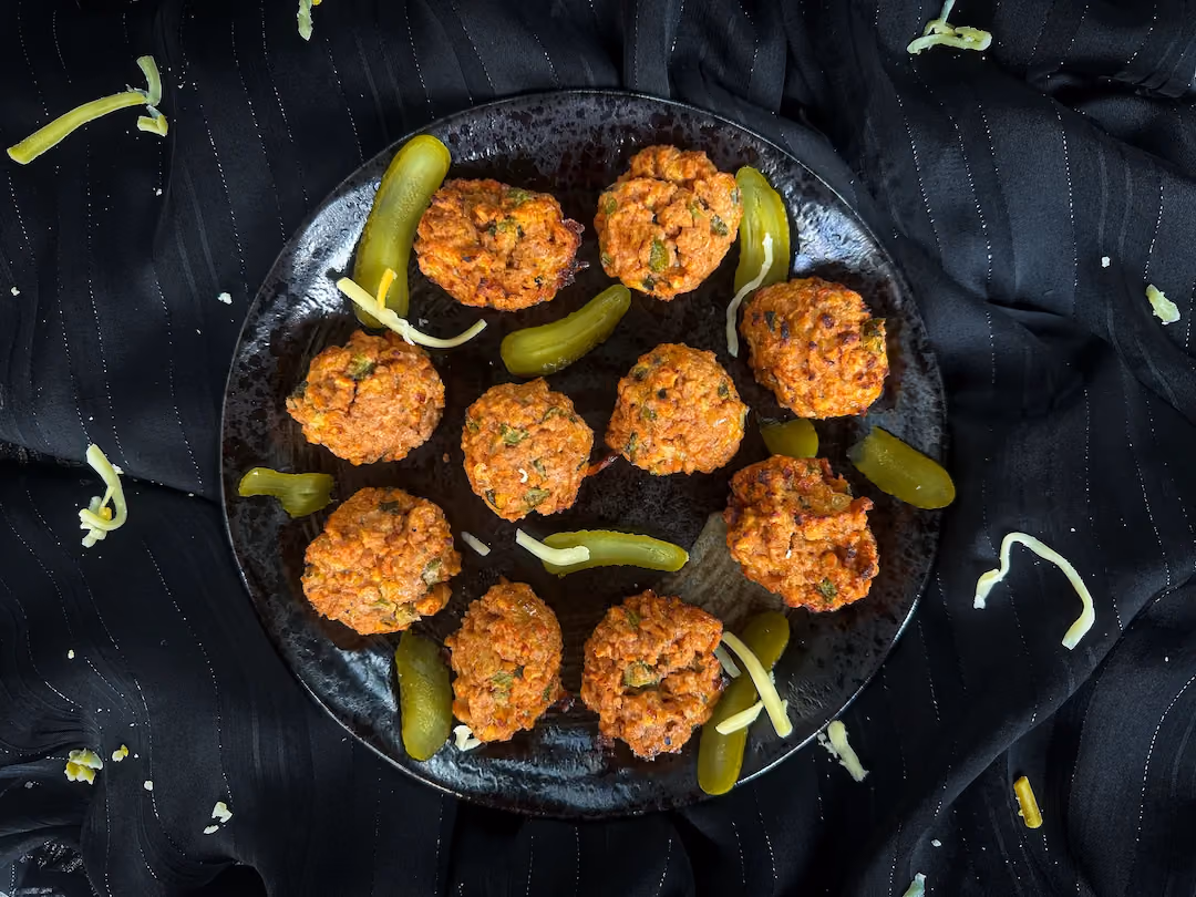 On a flat black plate are cheeseburger-style meatballs with pickle slices.