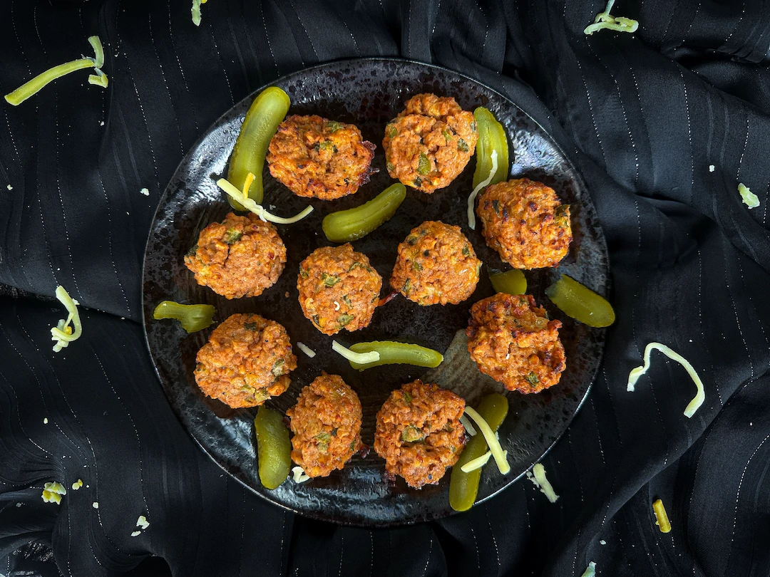 On a flat black plate are cheeseburger-style meatballs with pickle slices.