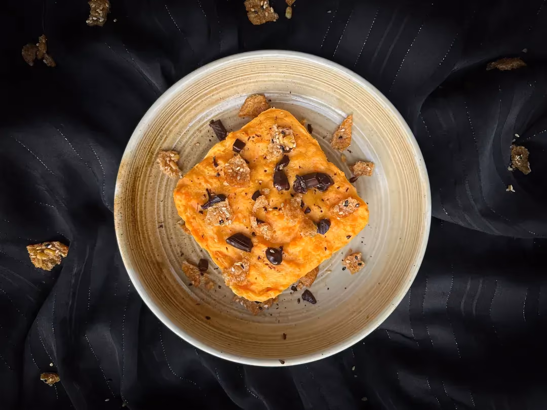Ce gâteau rond à la couleur orange est présenté sur une assiette noire déposée sur du feuillage et décoré autour d'ananas frais coupé grossièrement.