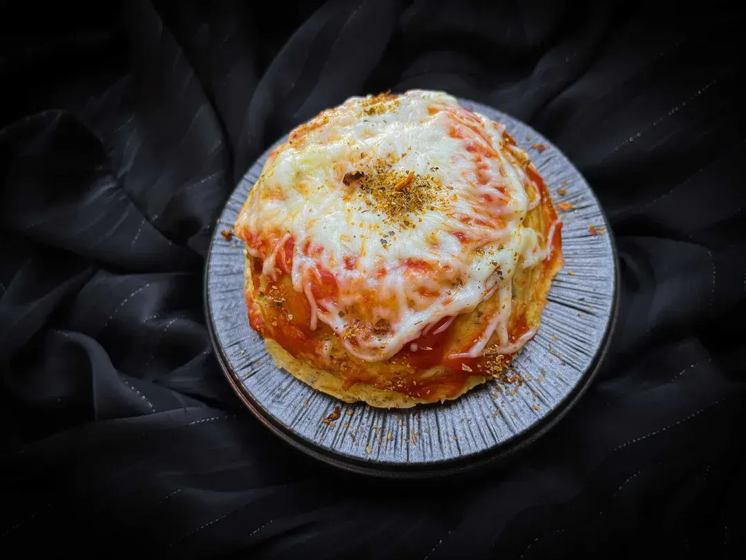 Dans un petit plat creux se trouve le Bowl cake façon pizza. Sur le dessus il y a du fromage râpé fondu.