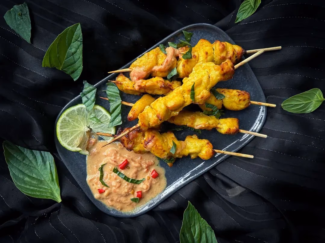 Une dizaine de petites brochettes de poulet à la sauce satay sont présentent sur un plat de couleur noir.
