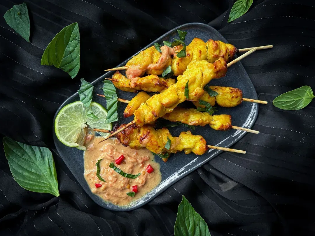 About ten small chicken satay skewers are arranged on a black plate.