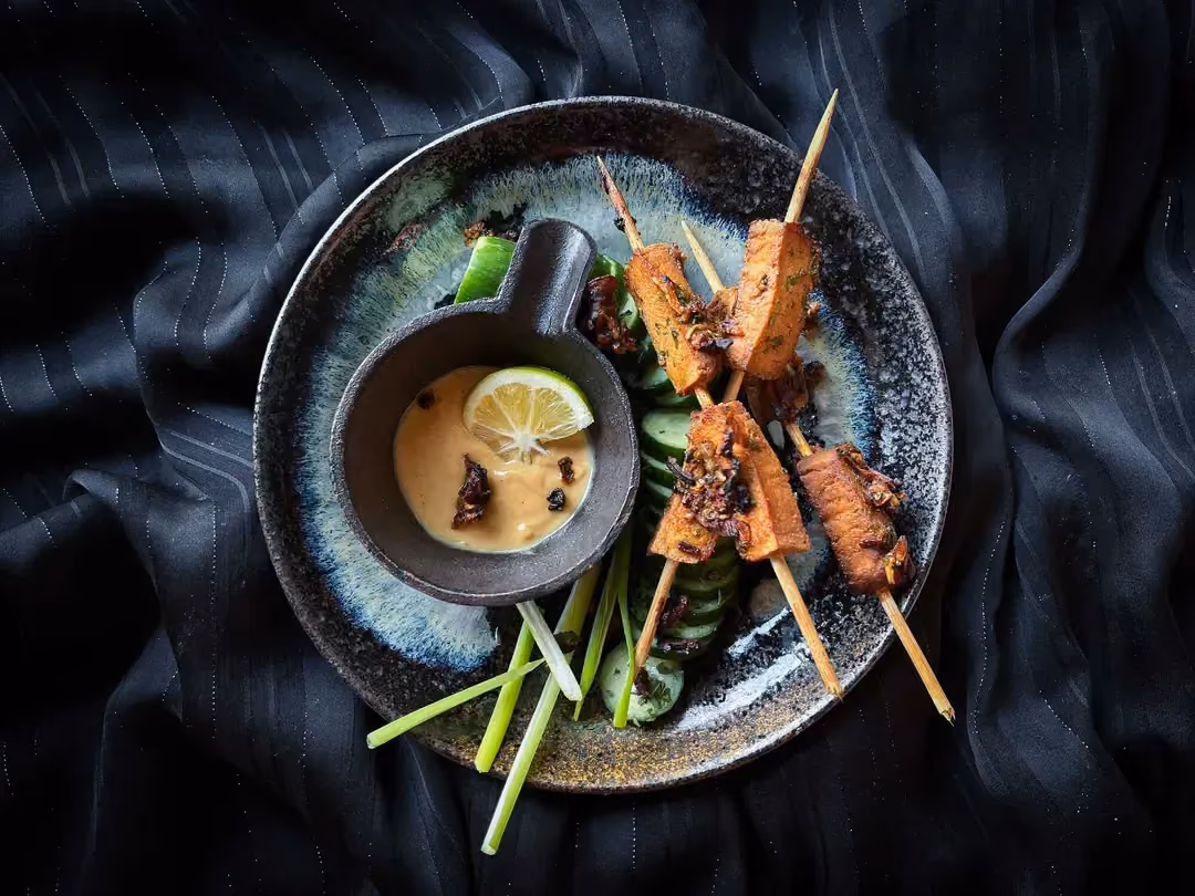 In a small blue bowl, there are three tofu skewers with satay sauce in a small ramekin.
