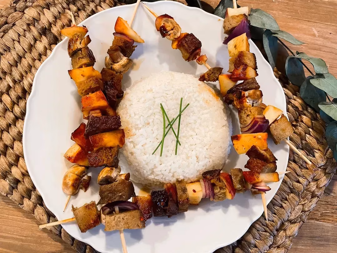 In the middle of a white plate is white rice, surrounded by five seitan skewers with apple and mushrooms.