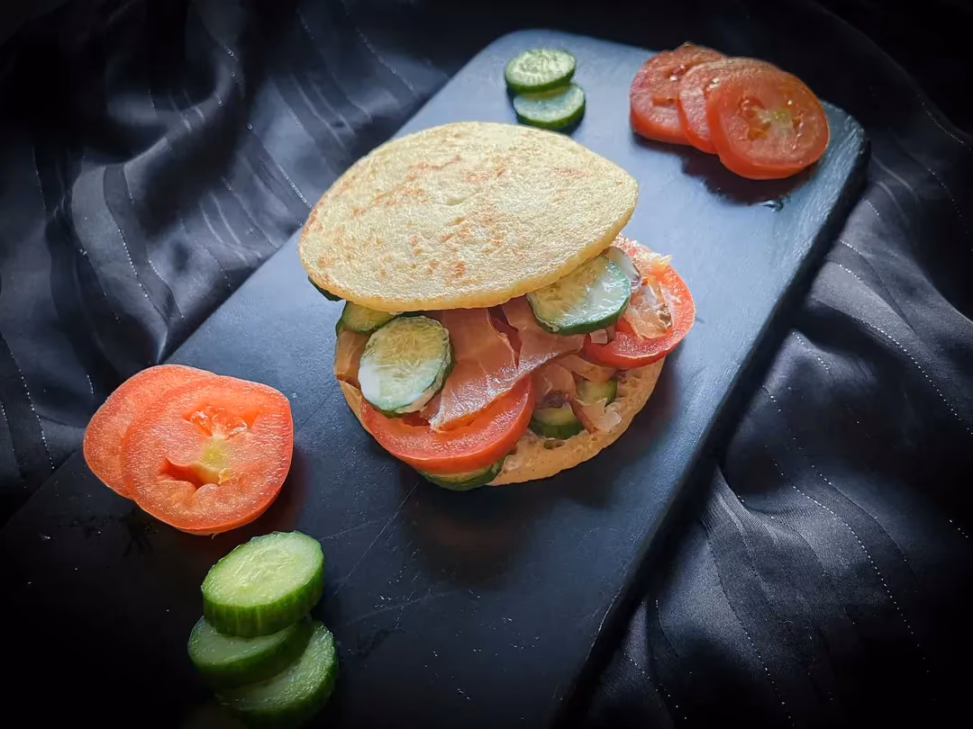 On a cutting board is a burger made with blinis, cucumber, tomatoes, smoked salmon, and blue cheese cream.