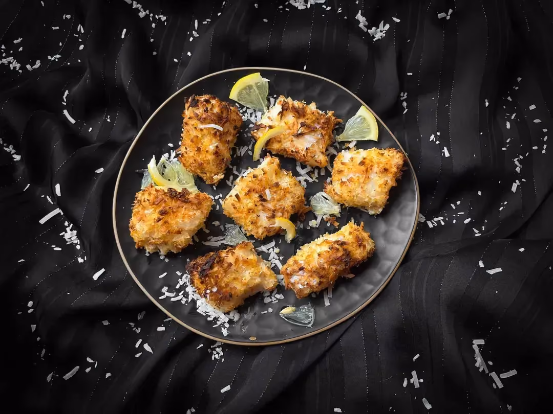 In a blue dish, there are seven pieces of cod breaded with coconut. Two thin slices of yellow lemon are present.