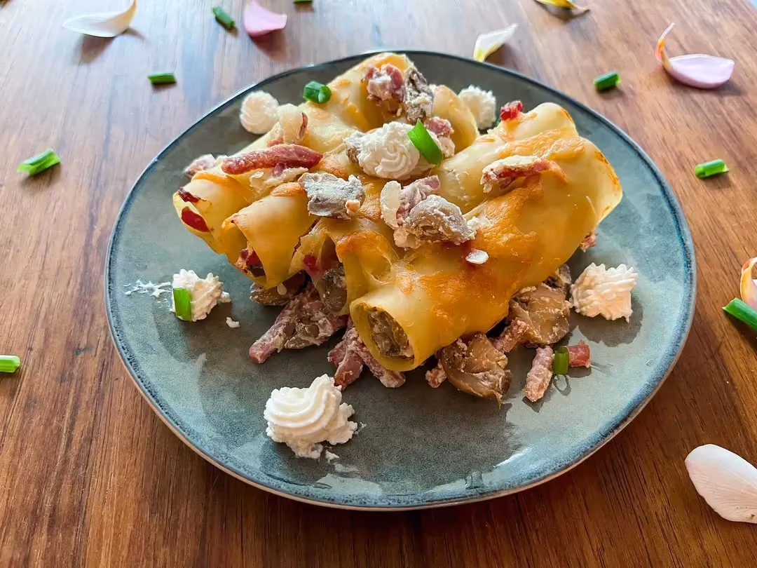 Déposée sur une table, une assiette de couleur bleu contient quatre gros cannelloni fourrés d'allumettes de bacon, champignon et ricotta.